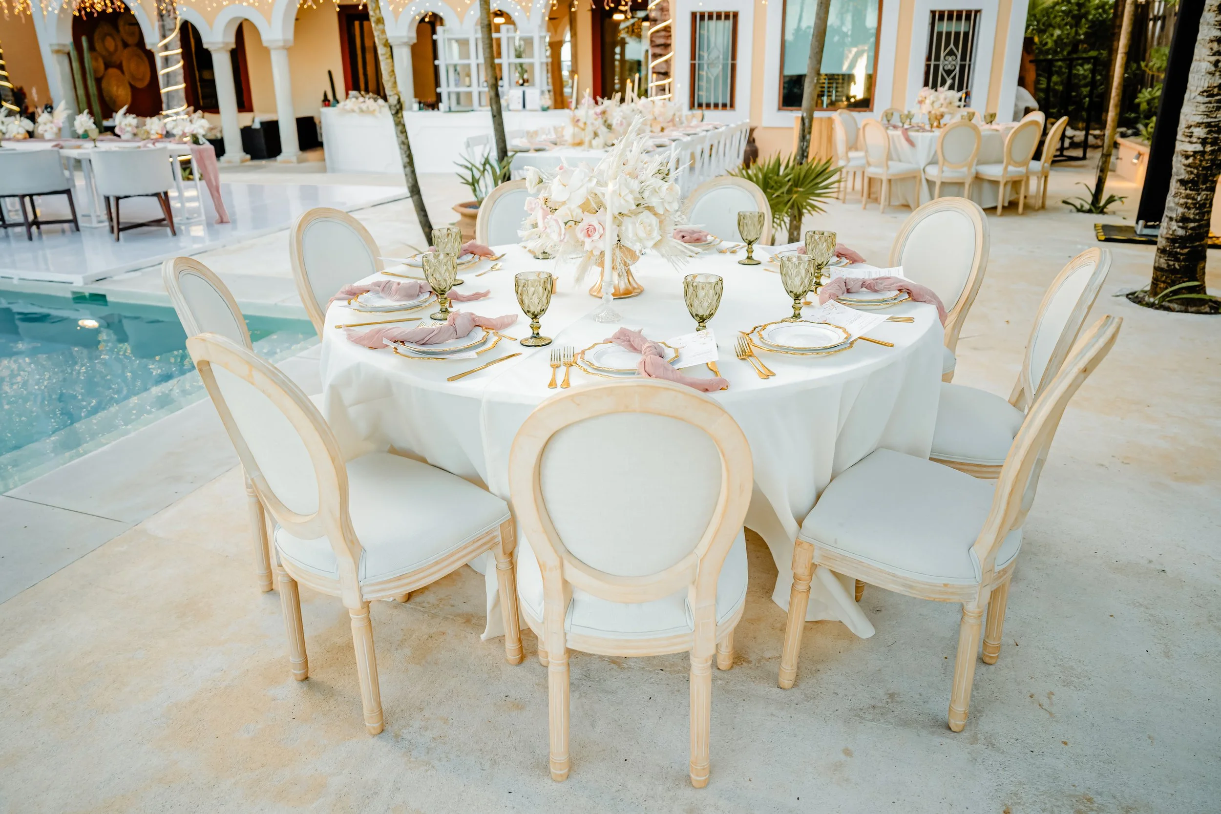 Round outdoor table set for a formal event with white tablecloth, gold-rimmed china, pink napkins, gold flatware, green tinted glassware, and a centerpiece of white and blush flowers, surrounded by cream-colored chairs, near a swimming pool and tropi