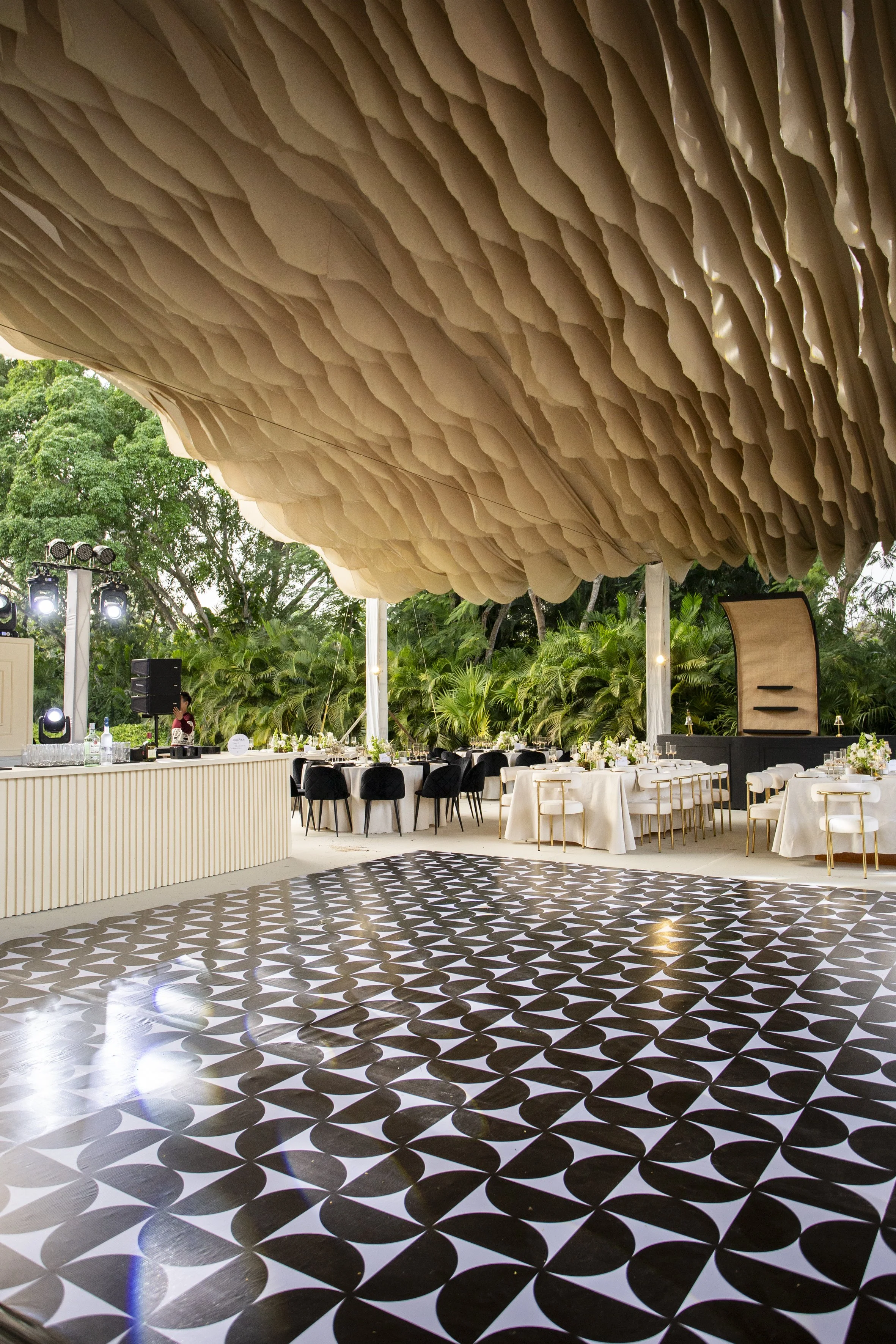 Elegant outdoor event venue with a decorated dance floor, round tables with white tablecloths and black or white chairs, lush green foliage in the background, and a large hanging decorative ceiling feature.
