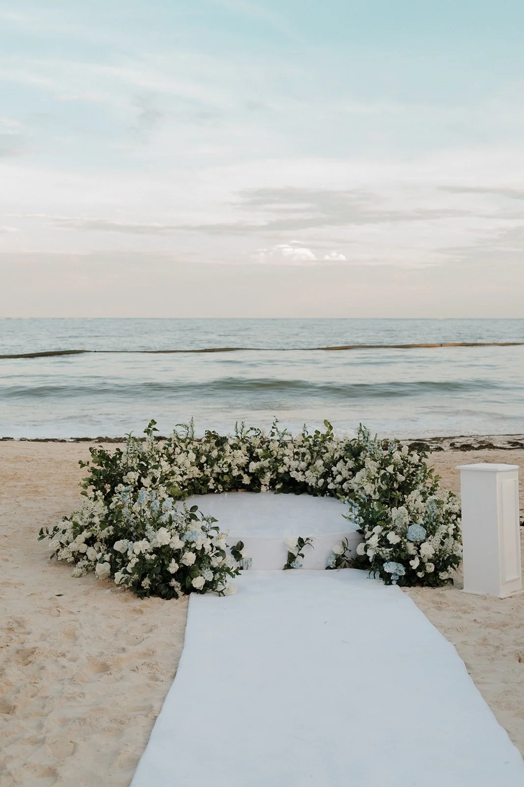 Wedding altar on a sandy beach with white floral arrangements and a white aisle runner, overlooking the ocean under a cloudy sky.