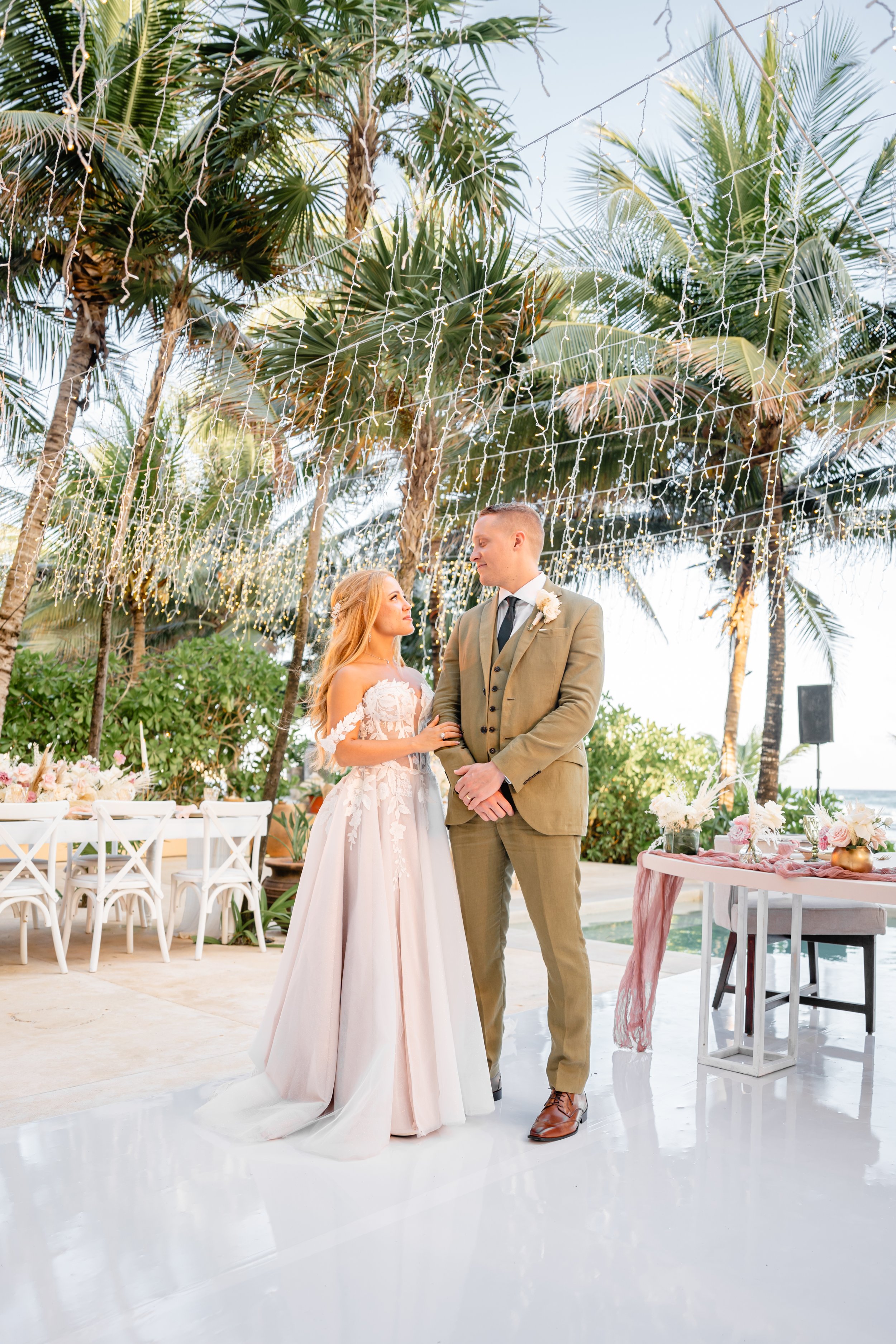 A bride and groom standing together at their outdoor wedding reception under string lights and palm trees, with seating and table decorations nearby.