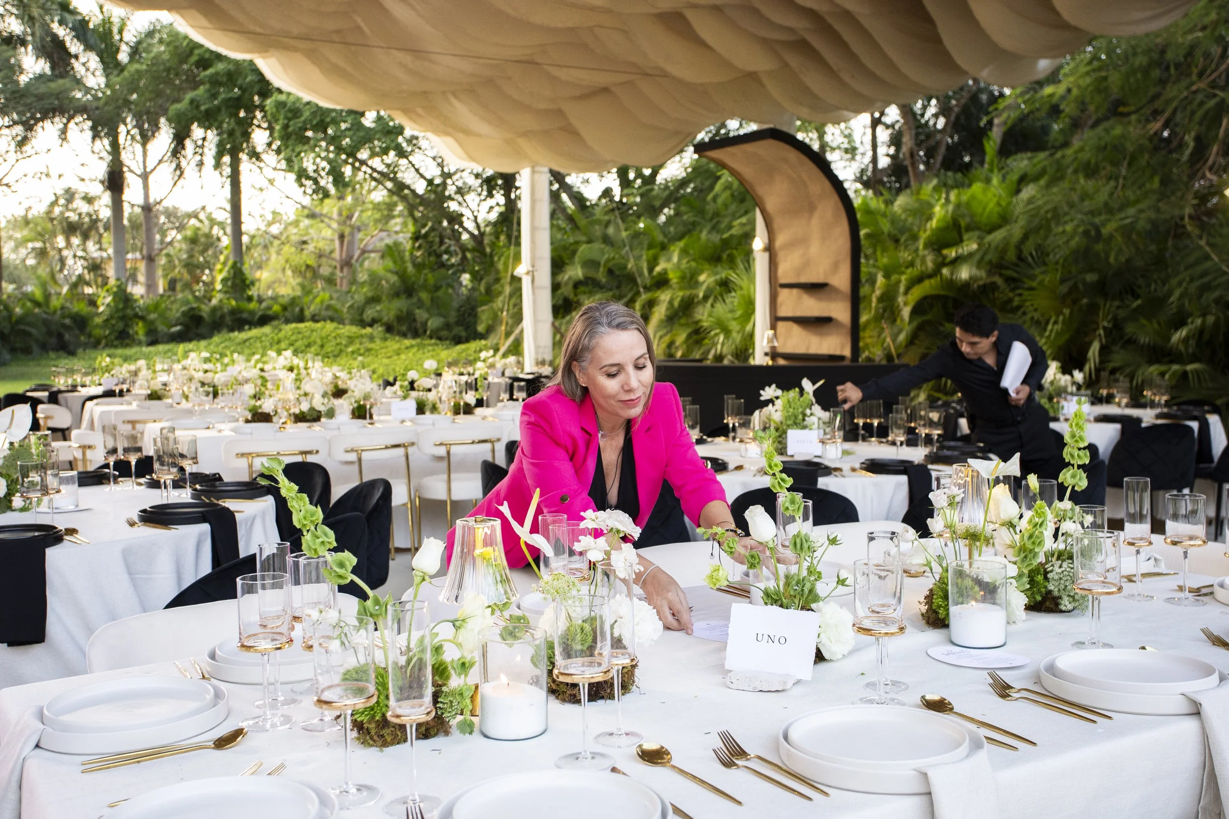 Alejandra Strazzulla, Creative Director at Scarlett Event Design in pink blazer sitting at an elegant outdoor banquet table with white tablecloth, floral centerpiece, and place settings, surrounded by lush greenery and trees at Chichi Suarez Hacienda