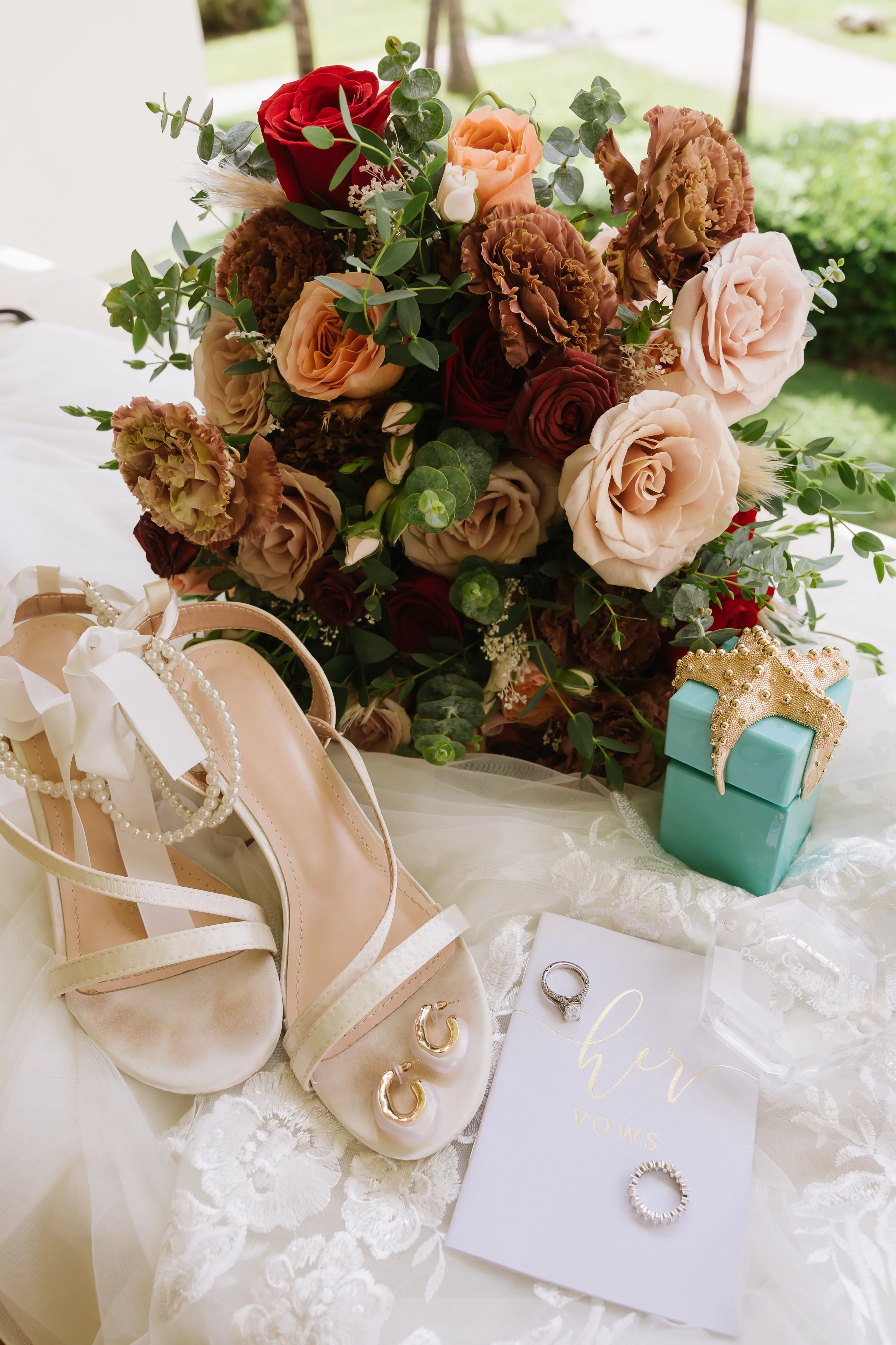 A bridal bouquet with roses and greenery, a pair of beige high-heeled shoes with pearl and ribbon decorations, gold hoop earrings, a ring, a vow card that says 'her vows', a turquoise gift box with a gold crown embellishment, and a clear plastic box,