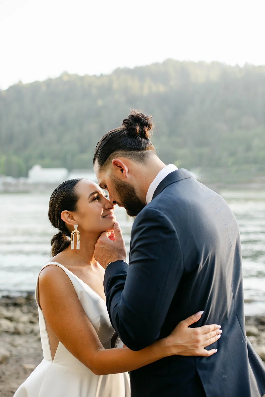 a couple embraces on the waterfront in portland, oregon at cathedral park. he holds her face and looks into her eyes lovingly. they are both indigenous and they are wearing wedding attire, a bride and groom.