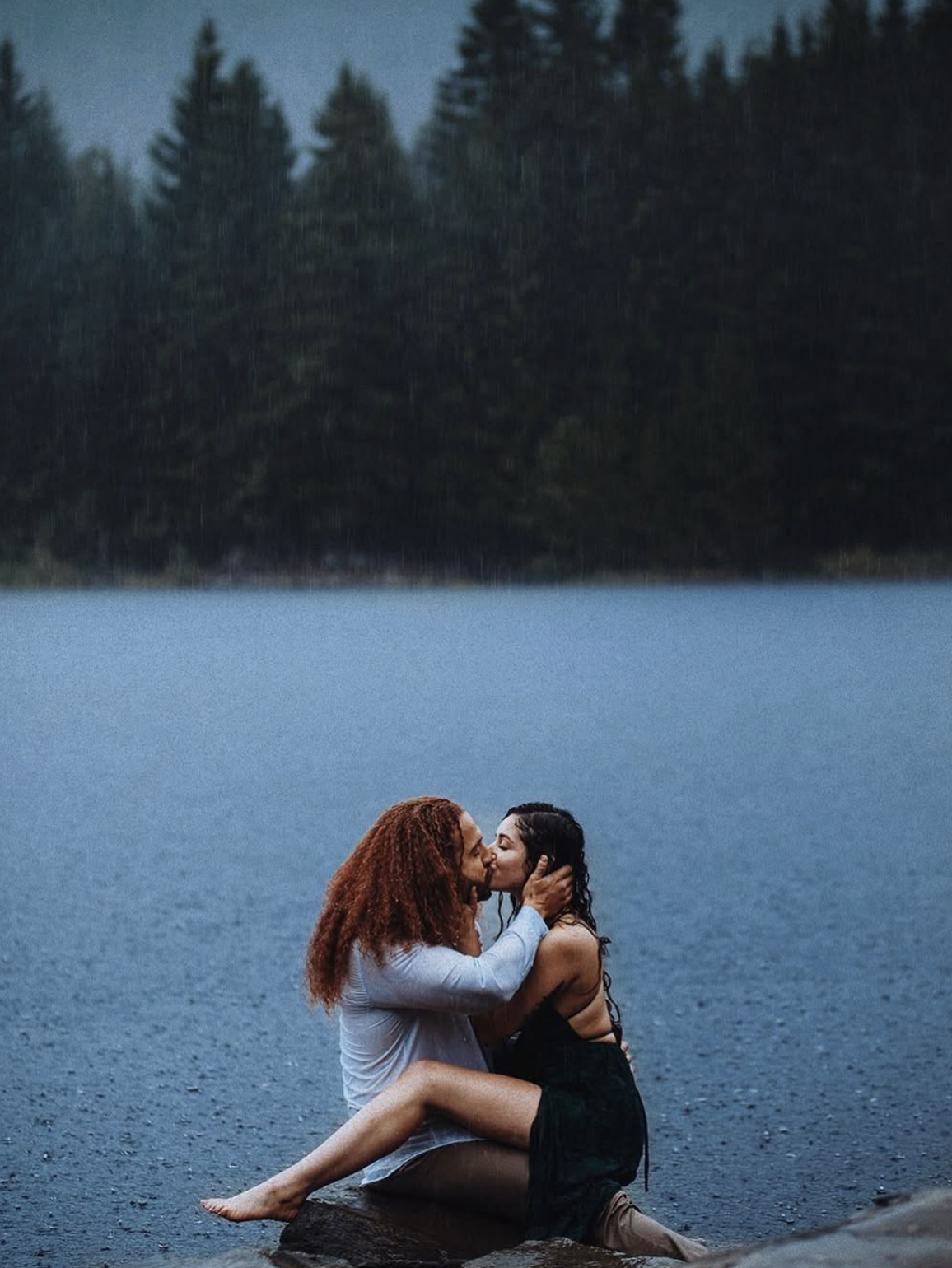 Rain soaked romantic couple sitting on a rock at Trillium Lake near Mount Hood Oregon on a moody rainy day, atmospheric Pacific Northwest engagement and elopement photography by Leah Flores of Wed n Wild, Portland Oregon