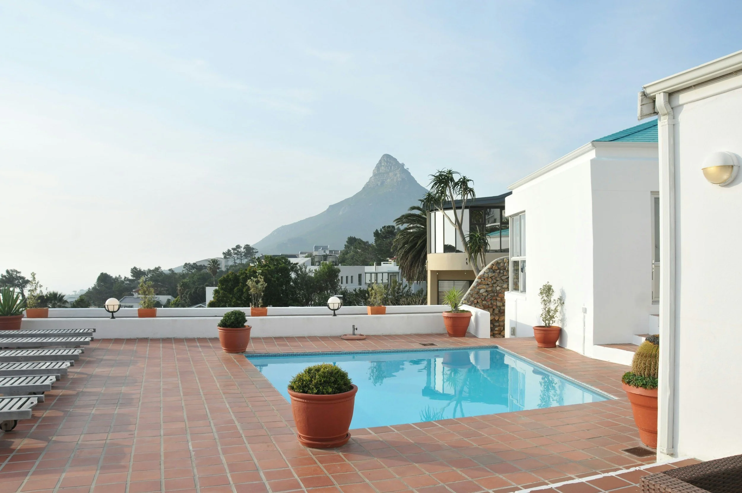 Outdoor rooftop pool area with terracotta tile flooring, potted plants, lounge chairs, white building, trees, distant mountains, and clear sky.