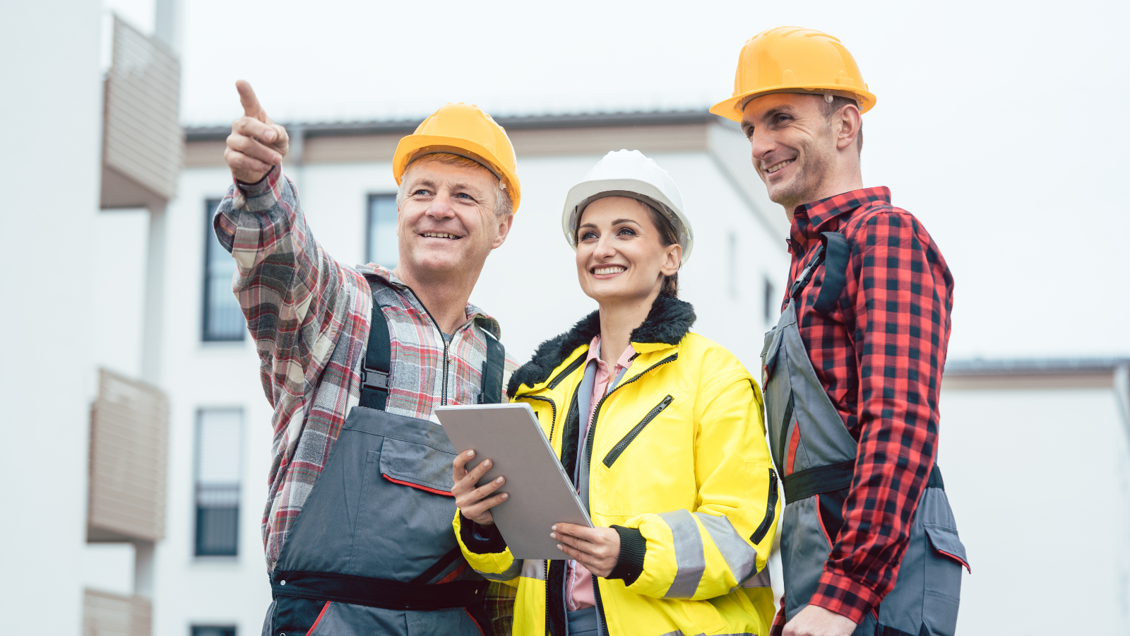 Three construction workers, two men and one woman, wearing safety helmets and reflective jackets, standing outdoors in front of residential buildings, discussing and smiling.