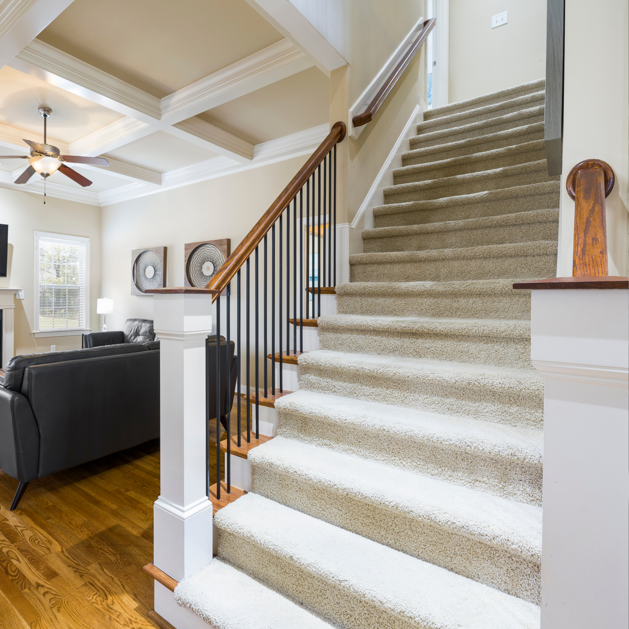 Interior view of a staircase with beige carpet, wooden handrail, and black metal balusters, next to a living room with black sofas, a window, and wall art.
