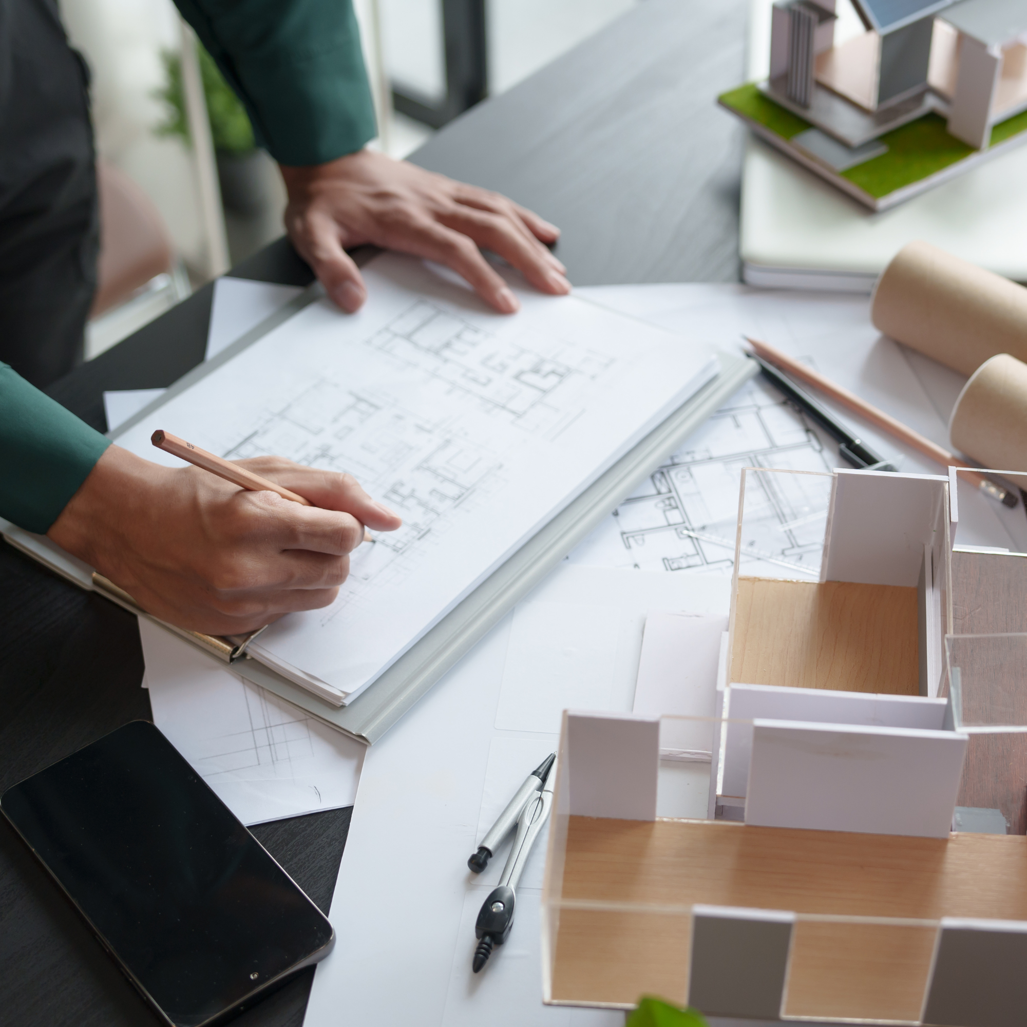 A person working on architectural blueprints at a desk, with model buildings, a pencil, pens, a smartphone, and rolled-up plans around.