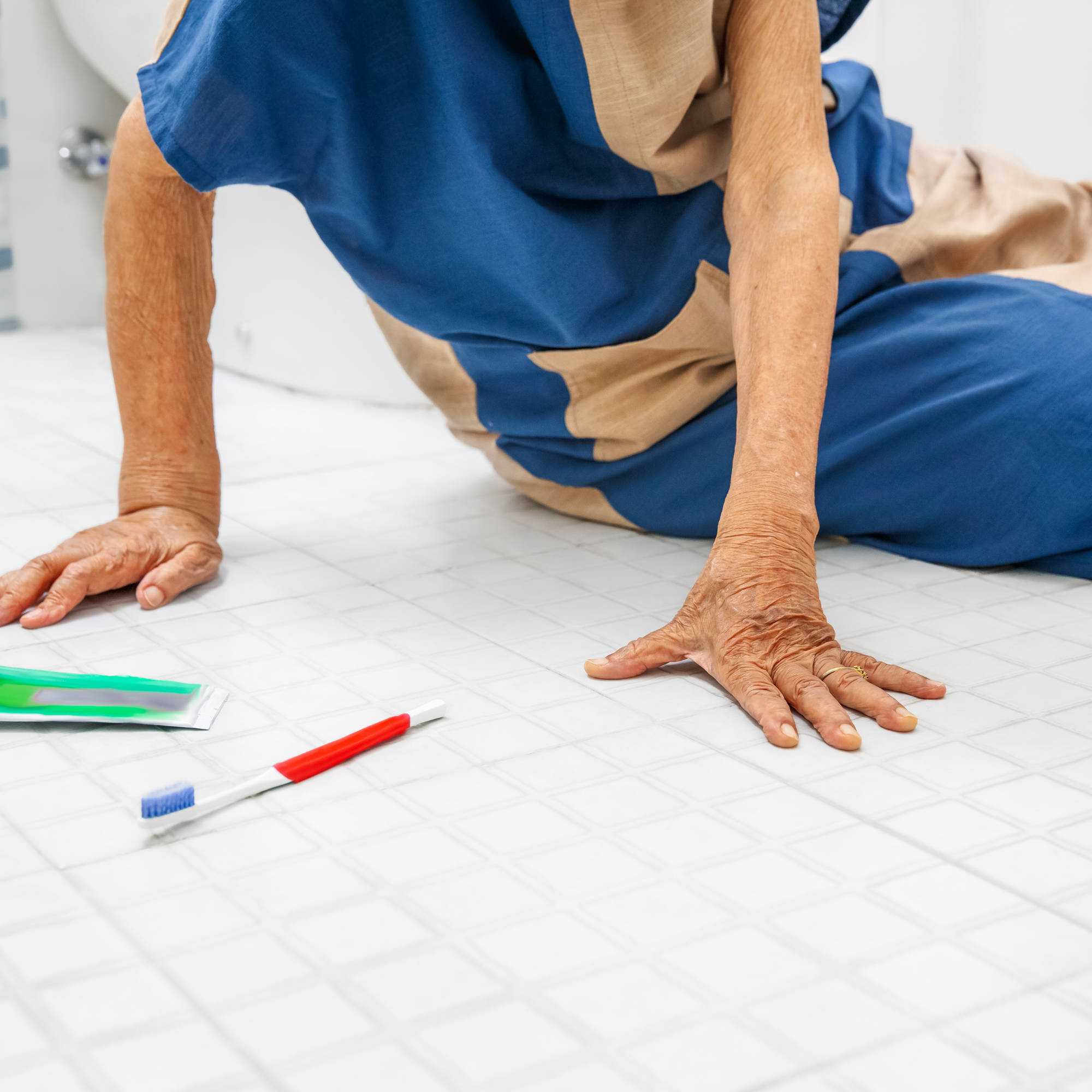 An elderly person kneels on a white tiled floor, supporting themselves with one hand, while a toothbrush and toothpaste lie nearby.