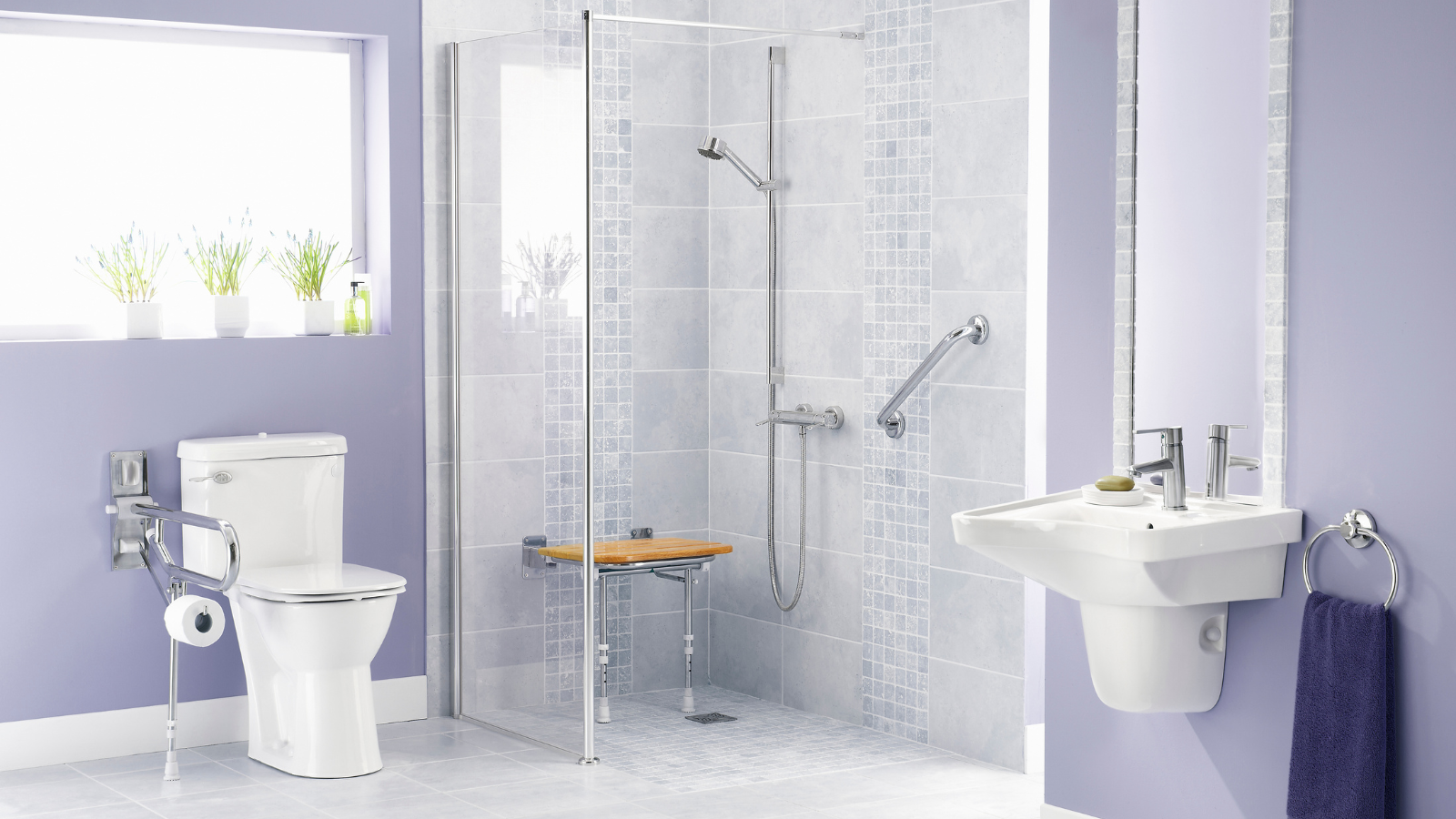 A bathroom featuring a toilet with a wall-mounted toilet paper holder, a walk-in shower with a glass door, gray tiled walls with decorative accents, a small wooden bench, a wall-mounted sink, and a purple towel hanging on a ring holder. There are three potted plants on a window sill with sunlight streaming in.