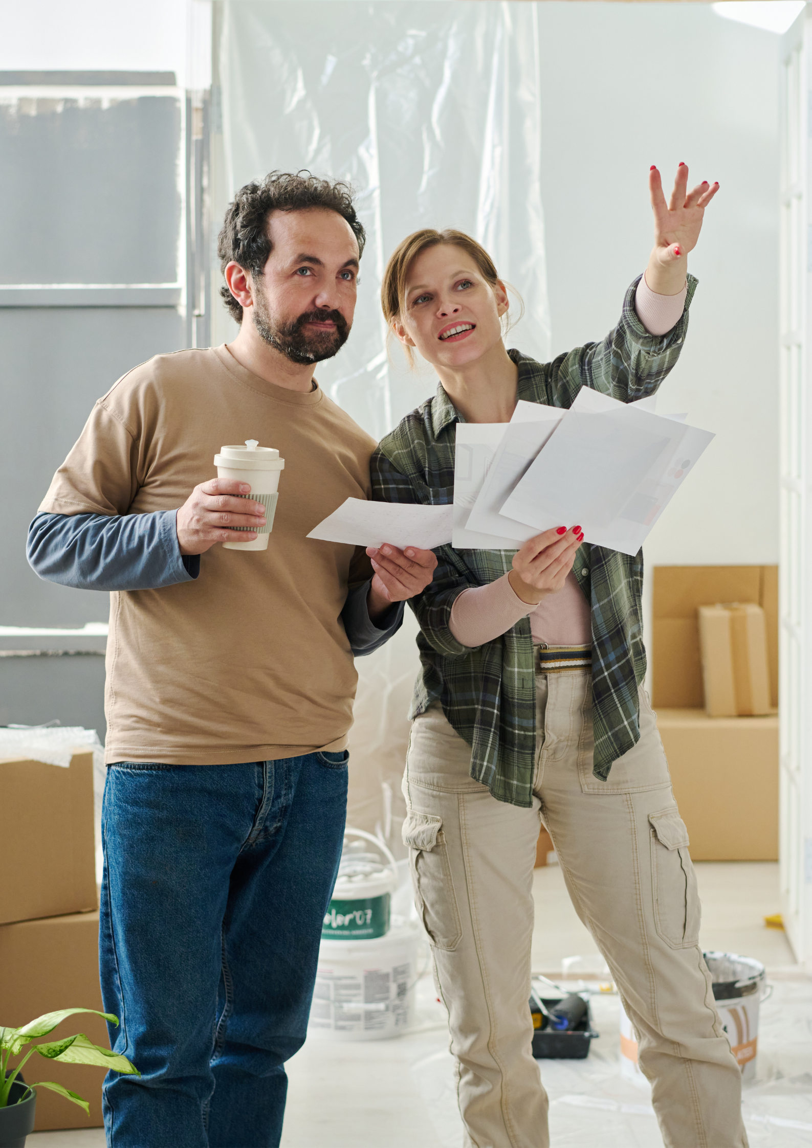 A man and woman discussing plans while holding papers, in a room with boxes and painting supplies, suggesting they are renovating or decorating a space.