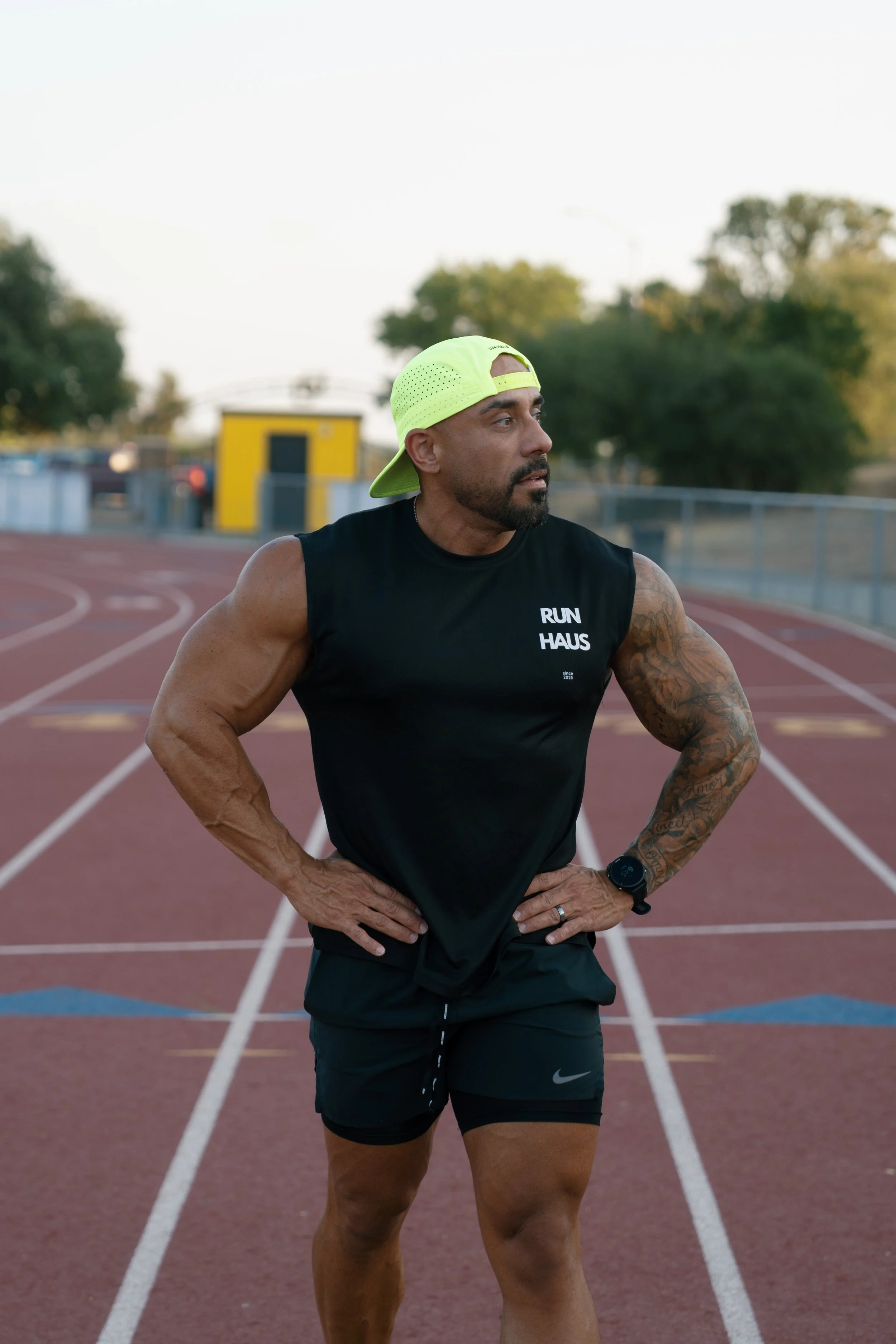 A man standing on a running track wearing a black sleeveless shirt with 'RUN HAUS' printed on it, black shorts, a black watch, and a neon yellow cap worn backwards, with his hands on his hips and looking to the side.
