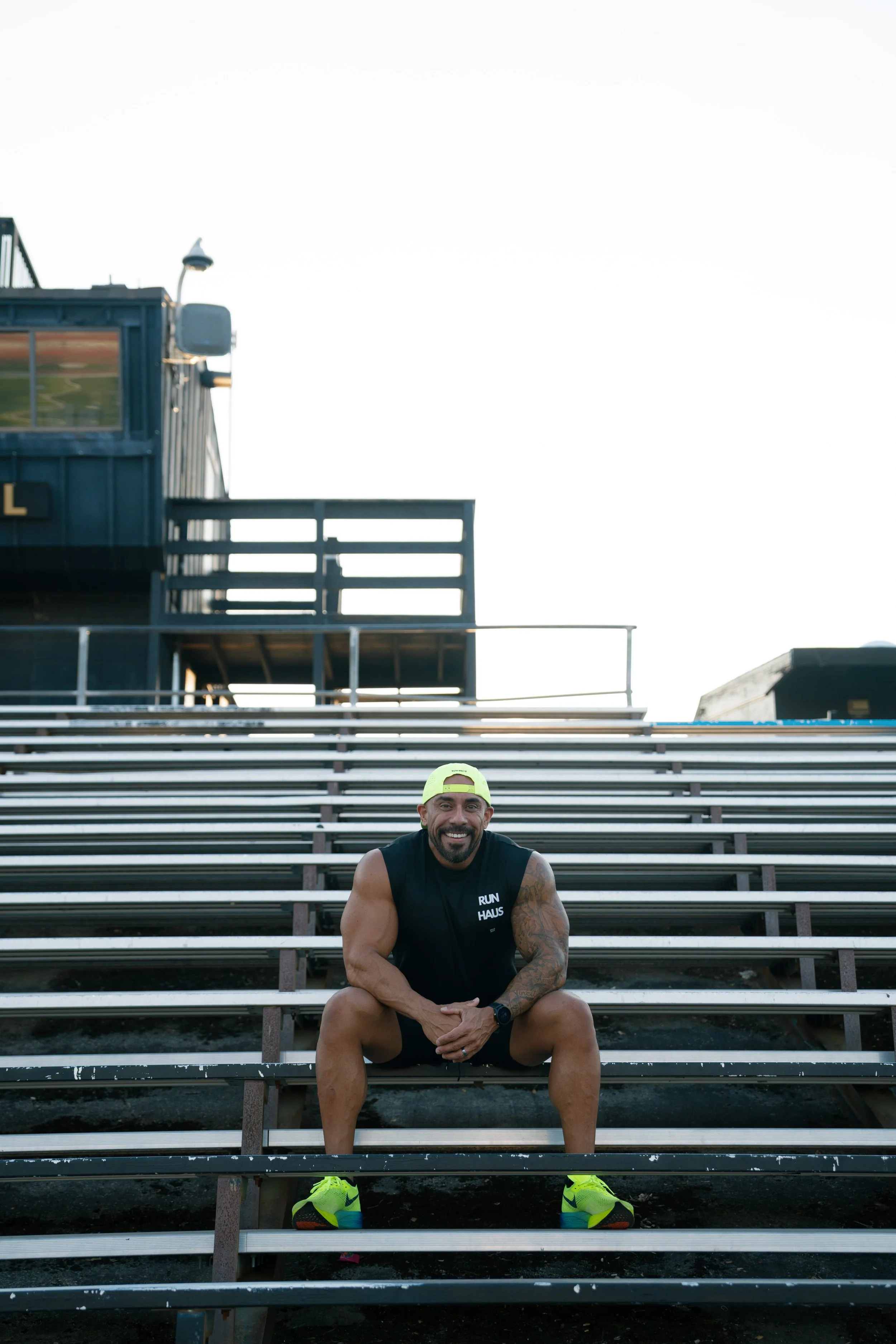 A man sitting on bleachers outdoors, smiling at the camera, wearing athletic clothing and bright yellow running shoes, with a building in the background.