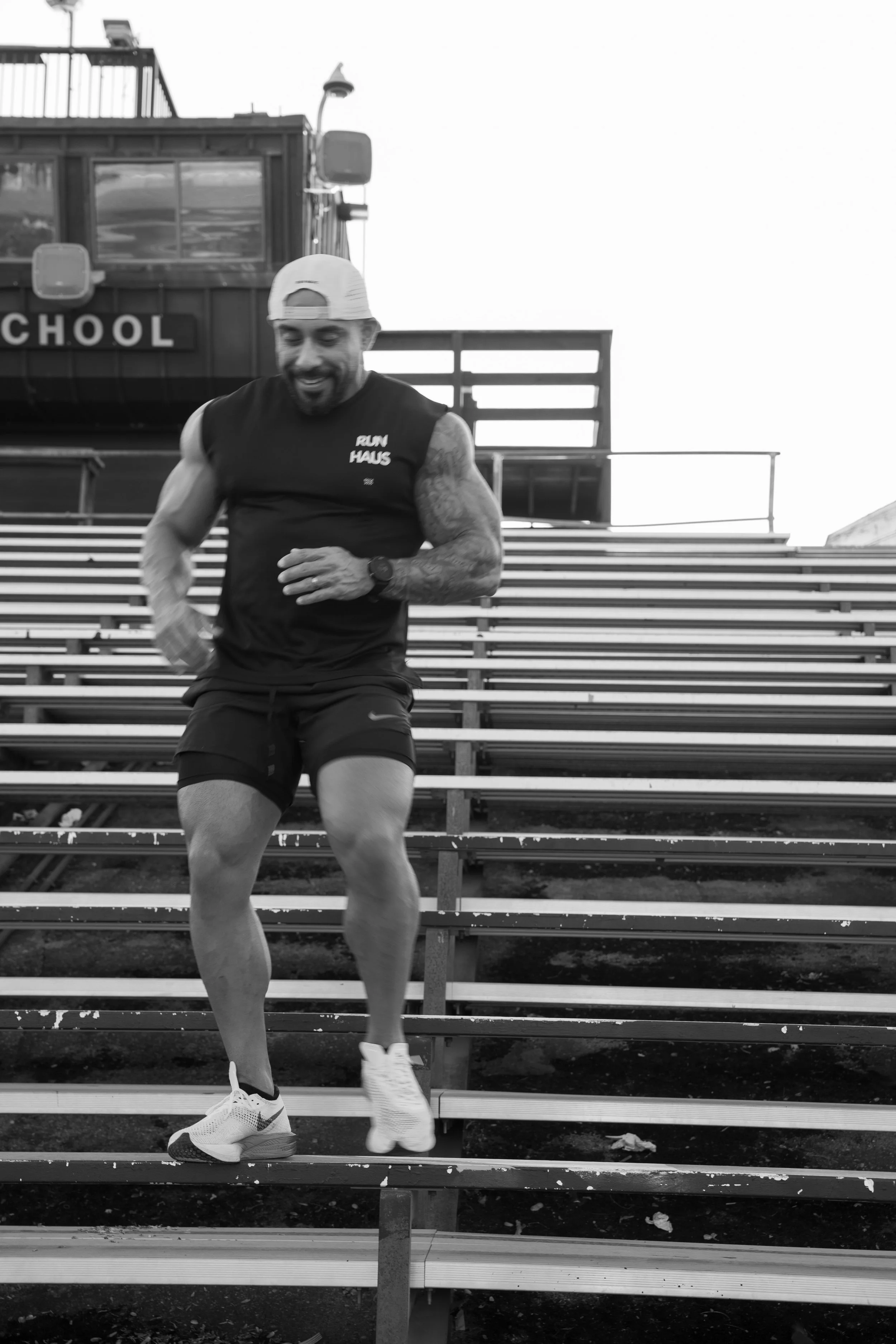 A man running down stadium bleachers with a smile, wearing a cap, sleeveless shirt, shorts, and running shoes.