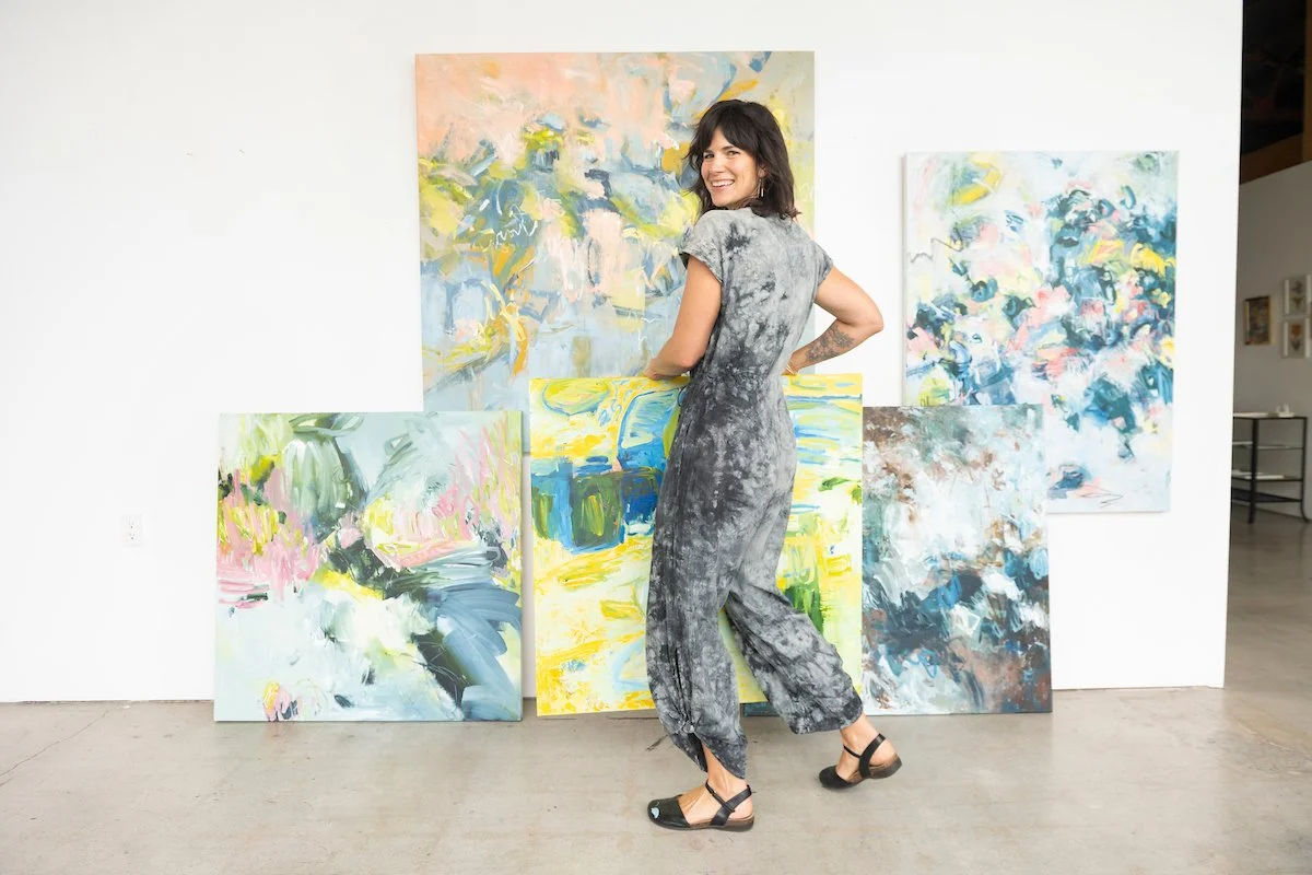 A woman in tie-dye jumpsuit standing in an art gallery with large abstract paintings on the wall behind her.