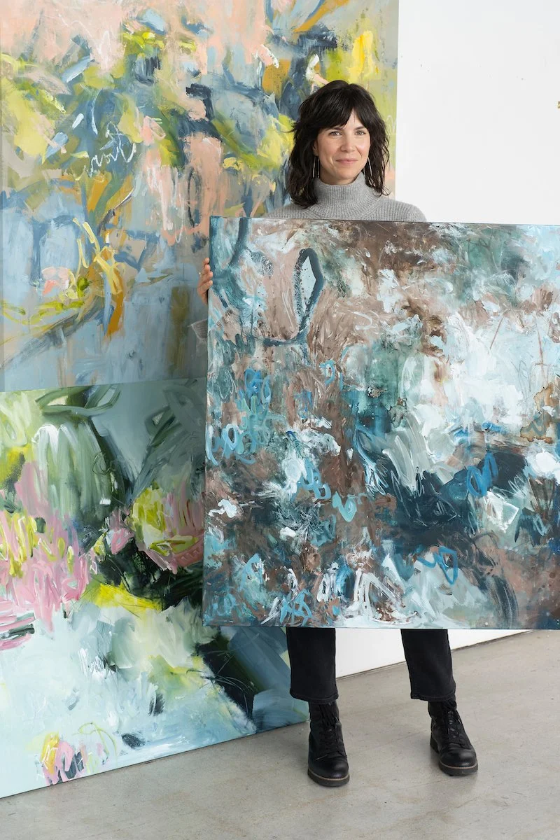 A woman standing in front of colorful abstract paintings, holding a large, abstract painting with blue, brown, and white brushstrokes.