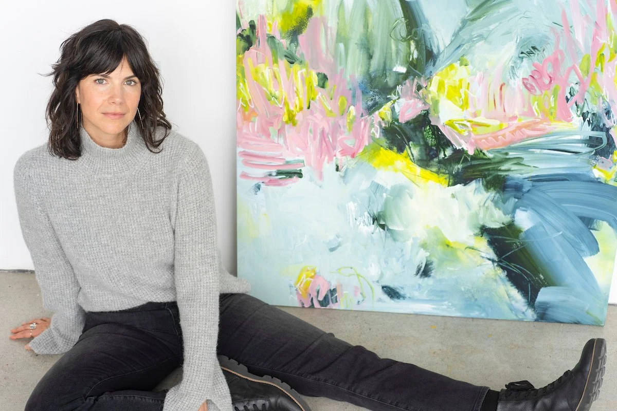 A woman sitting on the floor with her legs extended, leaning on her left hand, next to an abstract colorful painting. She has shoulder-length dark wavy hair, light skin, and is wearing a gray knit sweater, black jeans, and black shoes.