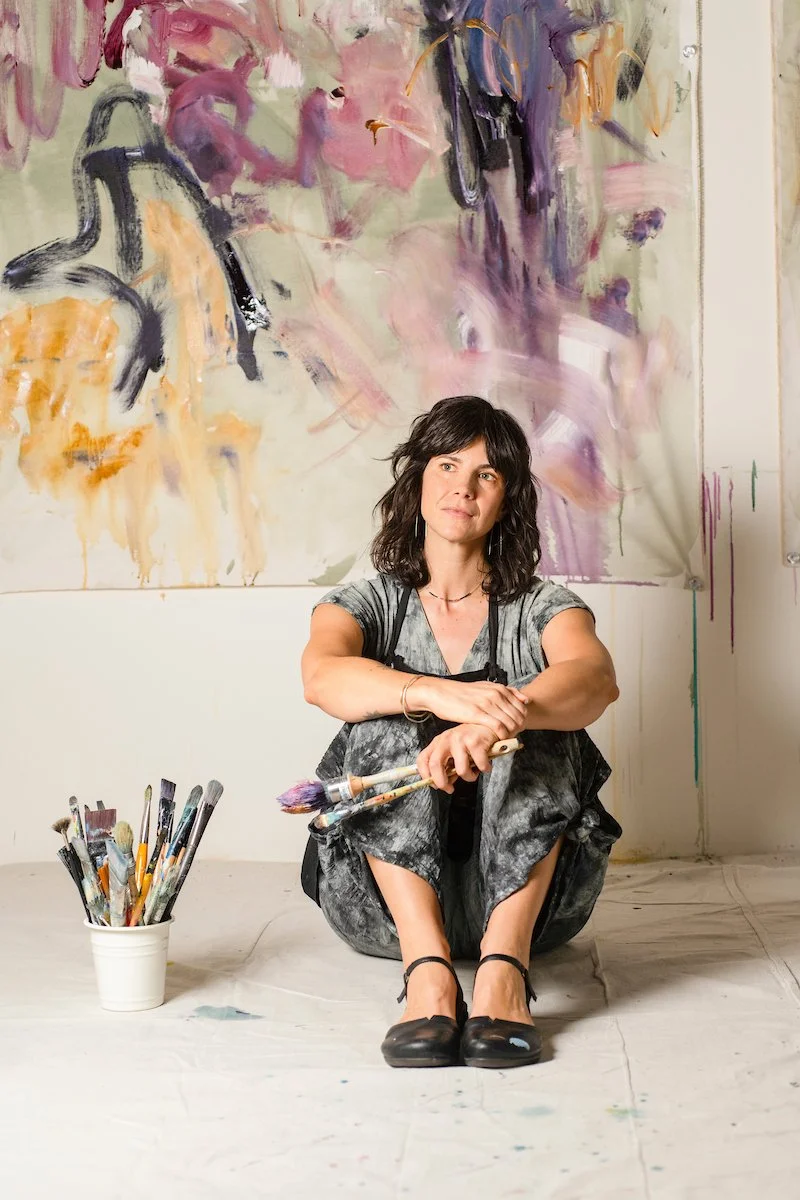 A woman sitting on the floor in front of a large, abstract, colorful painting with paintbrushes in her hands and a bucket of brushes beside her.