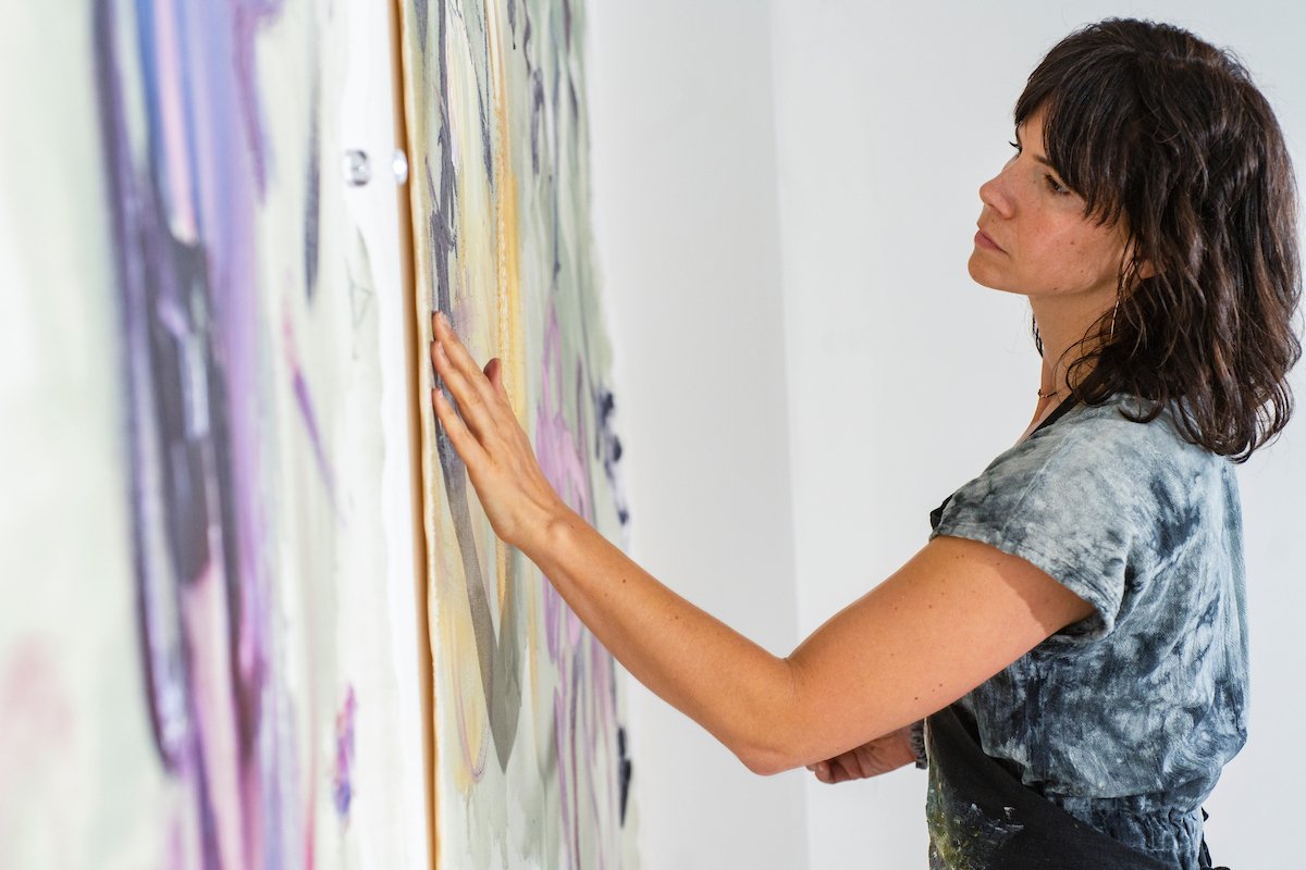 Woman with curly hair in a tie-dye shirt touches a colorful abstract painting on a gallery wall.