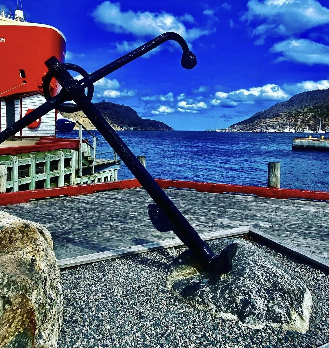 A large black anchor mounted on a rock at a waterfront, with a red ship and dock structures in the background. The scene features blue water and a partly cloudy sky.