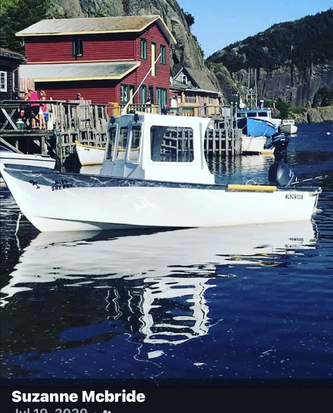 A small white fishing boat named 'Miss Sherry' is docked at a pier with multiple boats and colorful buildings with mountains in the background.