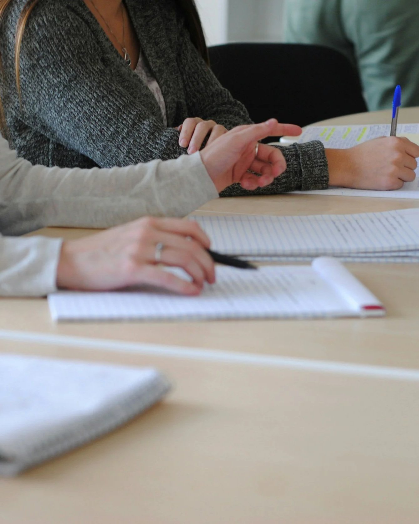 People sitting at a table, writing in notebooks with pens, in a classroom or meeting setting.