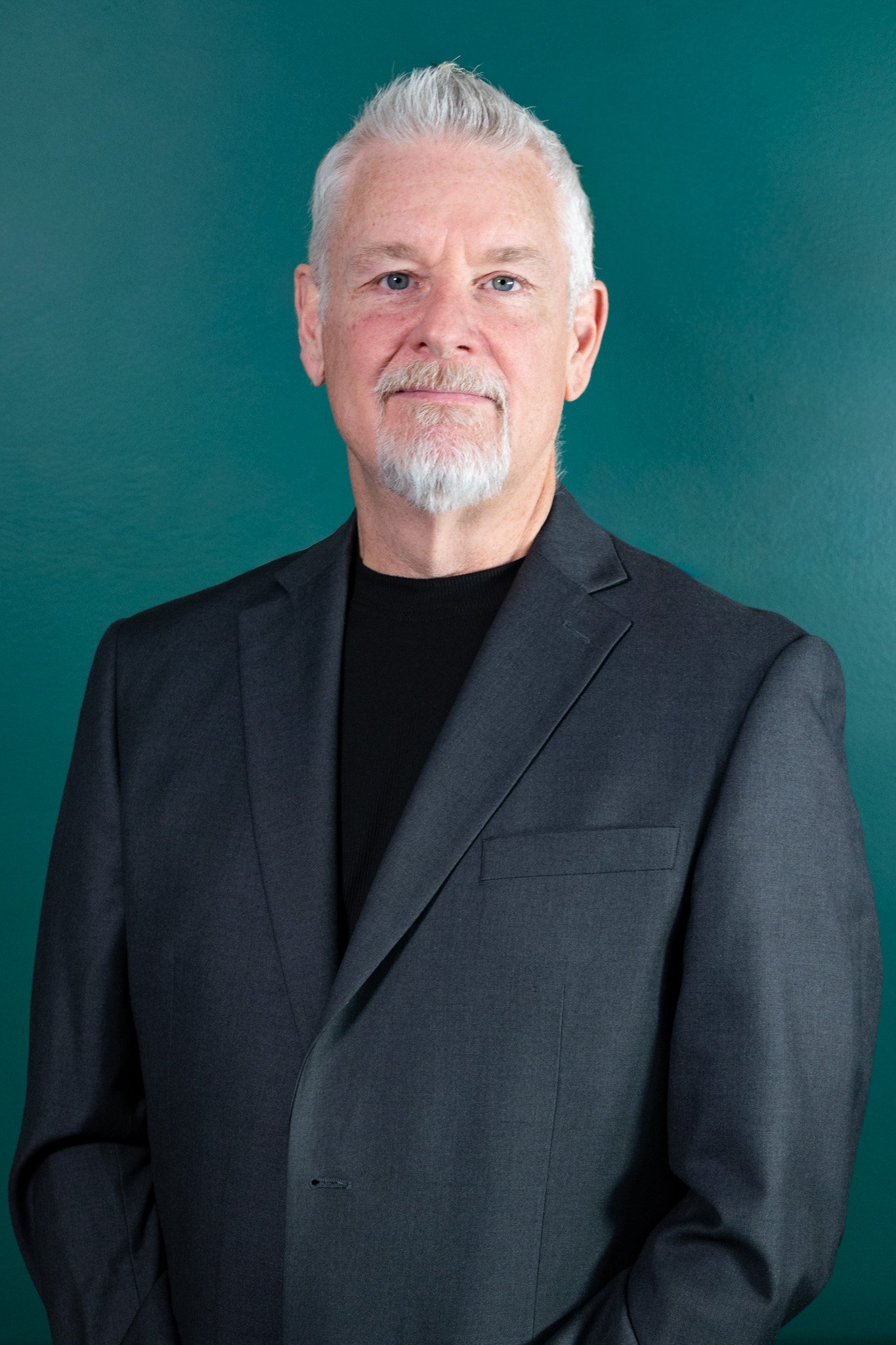 A portrait of Darin Voegeli, wearing a black blazer and black shirt, standing against a teal background.