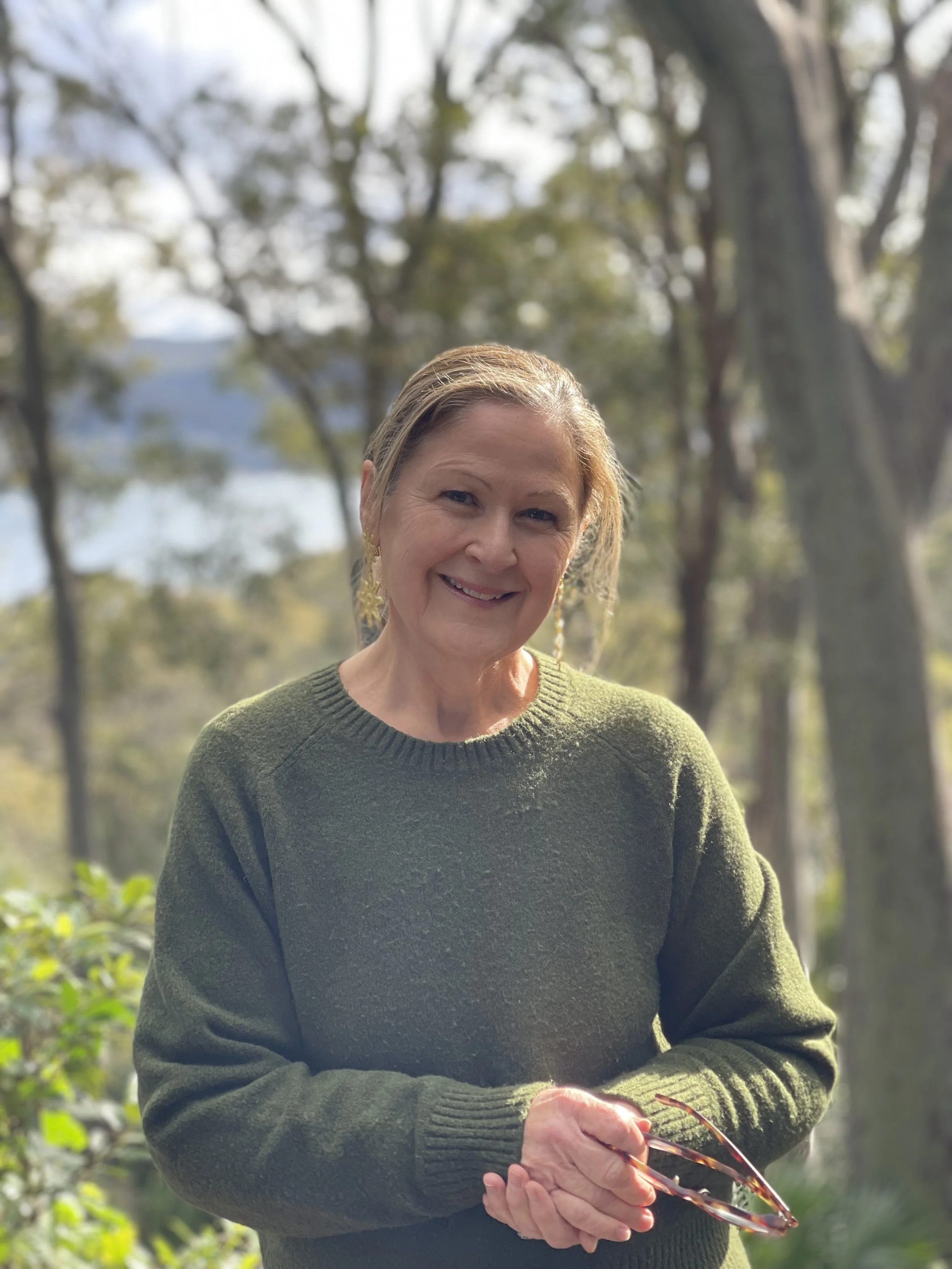 Smiling woman outdoors in a green sweater holding sunglasses, surrounded by trees and sunlight.