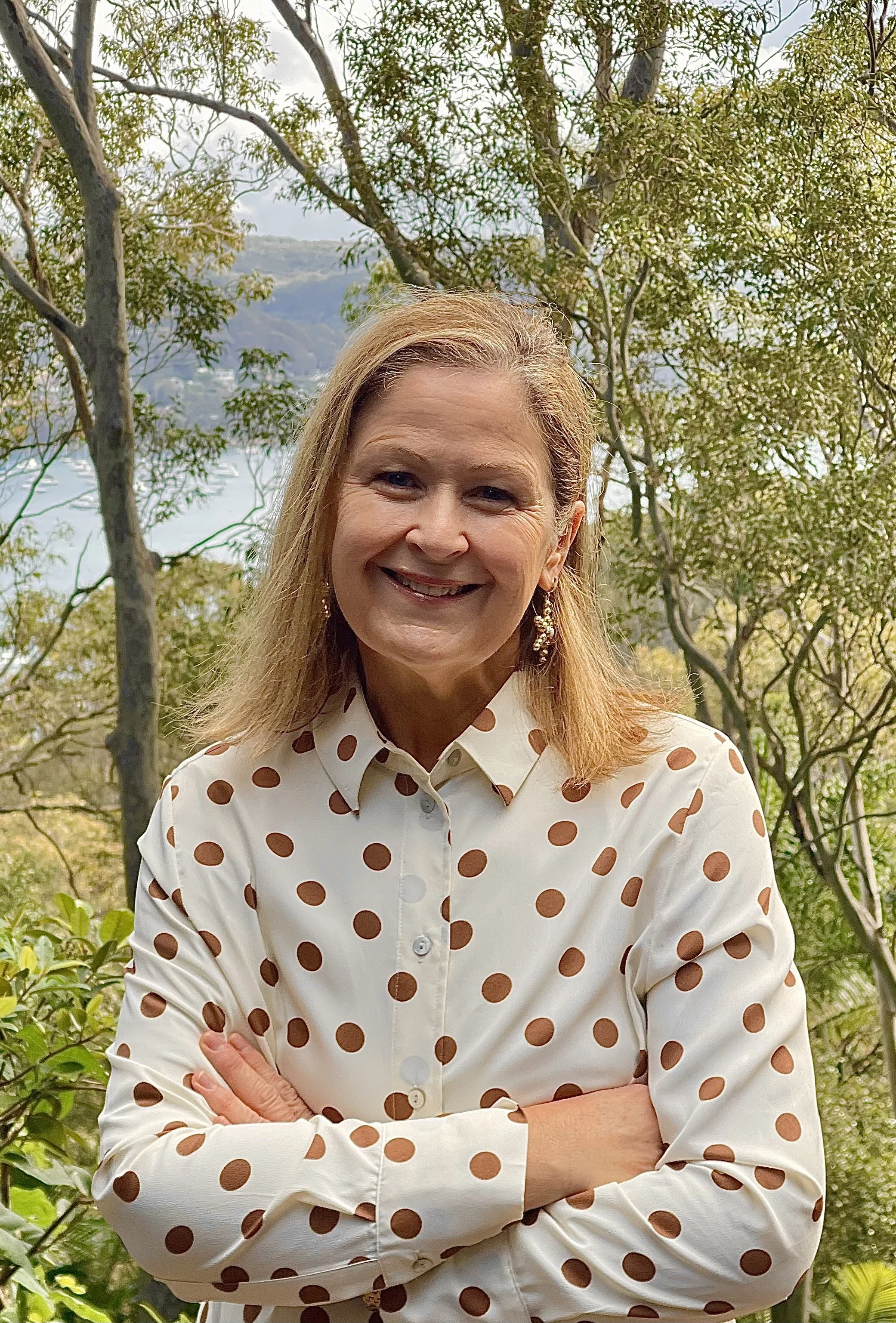 A woman smiling outdoors among trees and greenery, wearing a white shirt with large brown polka dots and earrings.