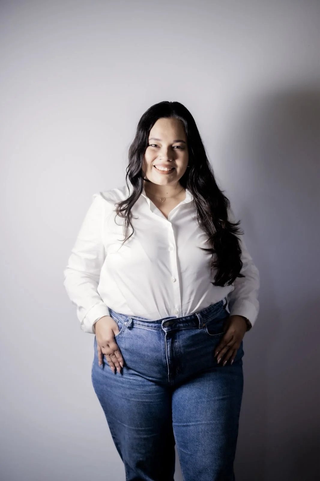 A young woman with long dark hair wearing a white button-up shirt and blue jeans, standing against a plain light gray background, smiling and posing with her hands in her pockets.