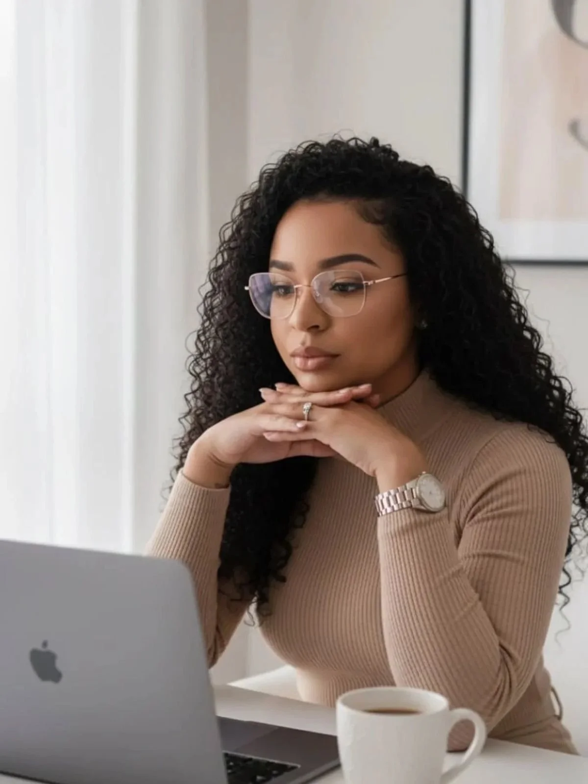A woman with curly hair and glasses sitting at a desk, looking at a laptop, with a white mug and a wristwatch.