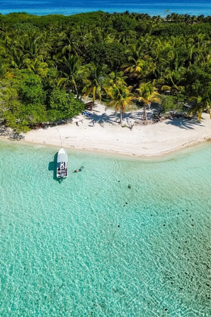Aerial view of a tropical beach with white sand, clear turquoise water, and dense green palm trees near the shoreline. A boat is anchored in shallow water, with people swimming nearby.