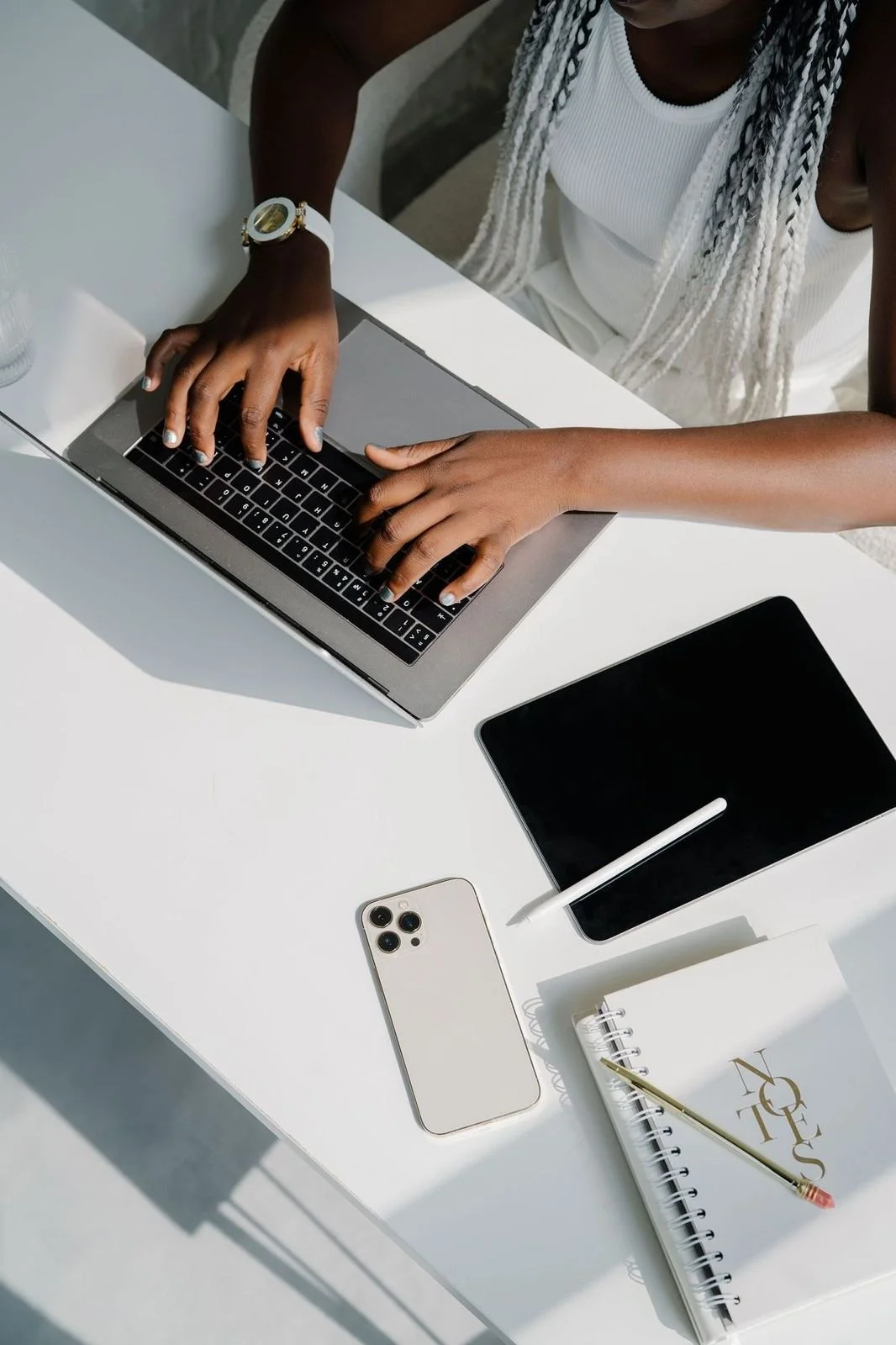 A woman working on a silver laptop at a white desk, with an iPhone, a graphics tablet, a notebook, and a pen nearby.