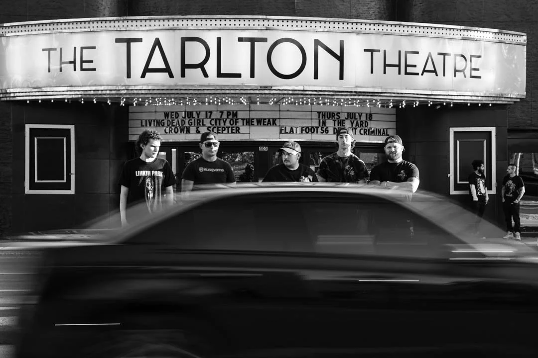 A black and white photo of the Tarlton Theatre marquee with five guys, Crown and Scepter, standing in front of it, and a blurred car passing by in the foreground.