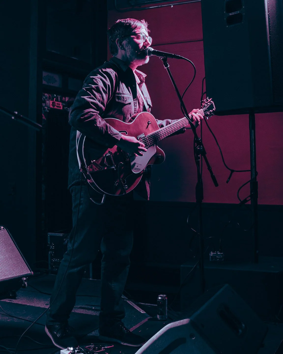 A man with glasses and a beard playing an acoustic guitar and singing into a microphone on stage, with purple and pink lighting.