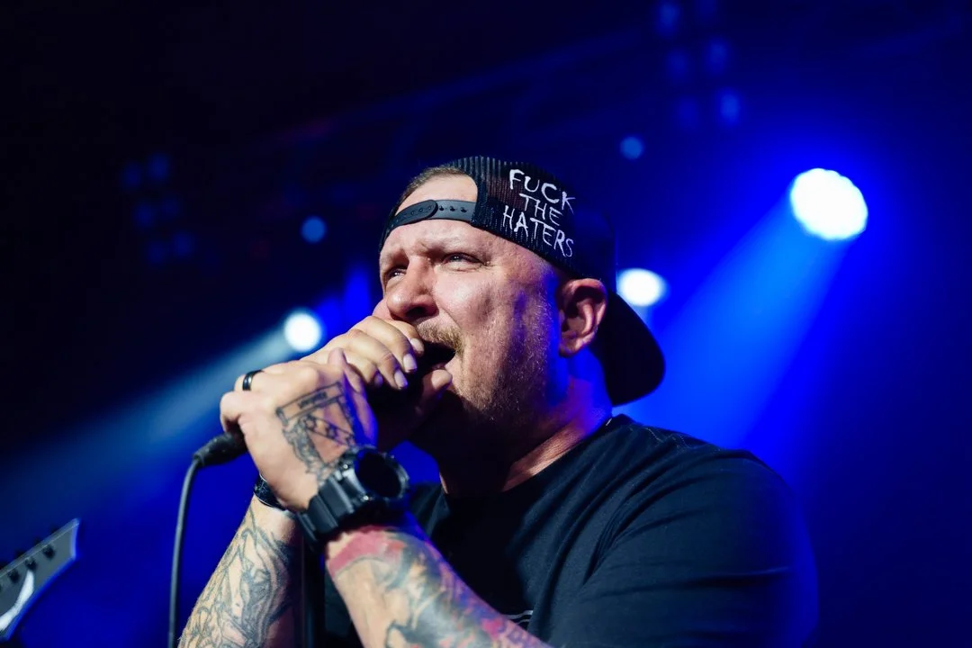 Male singer wearing a backward cap with 'F**k the Haters' written on it, passionately singing into a microphone on stage with blue and purple lighting.