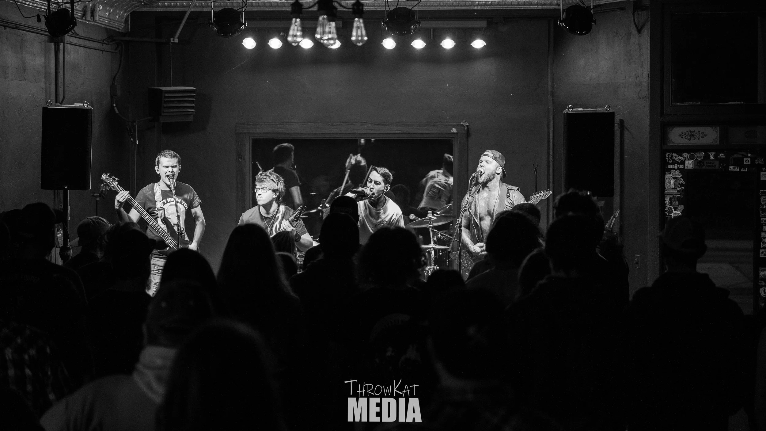 A band performs live on stage at a small music venue. The black and white photo shows four musicians with guitars and drums, facing an audience with heads silhouetted in the foreground. Stage lights hang above.