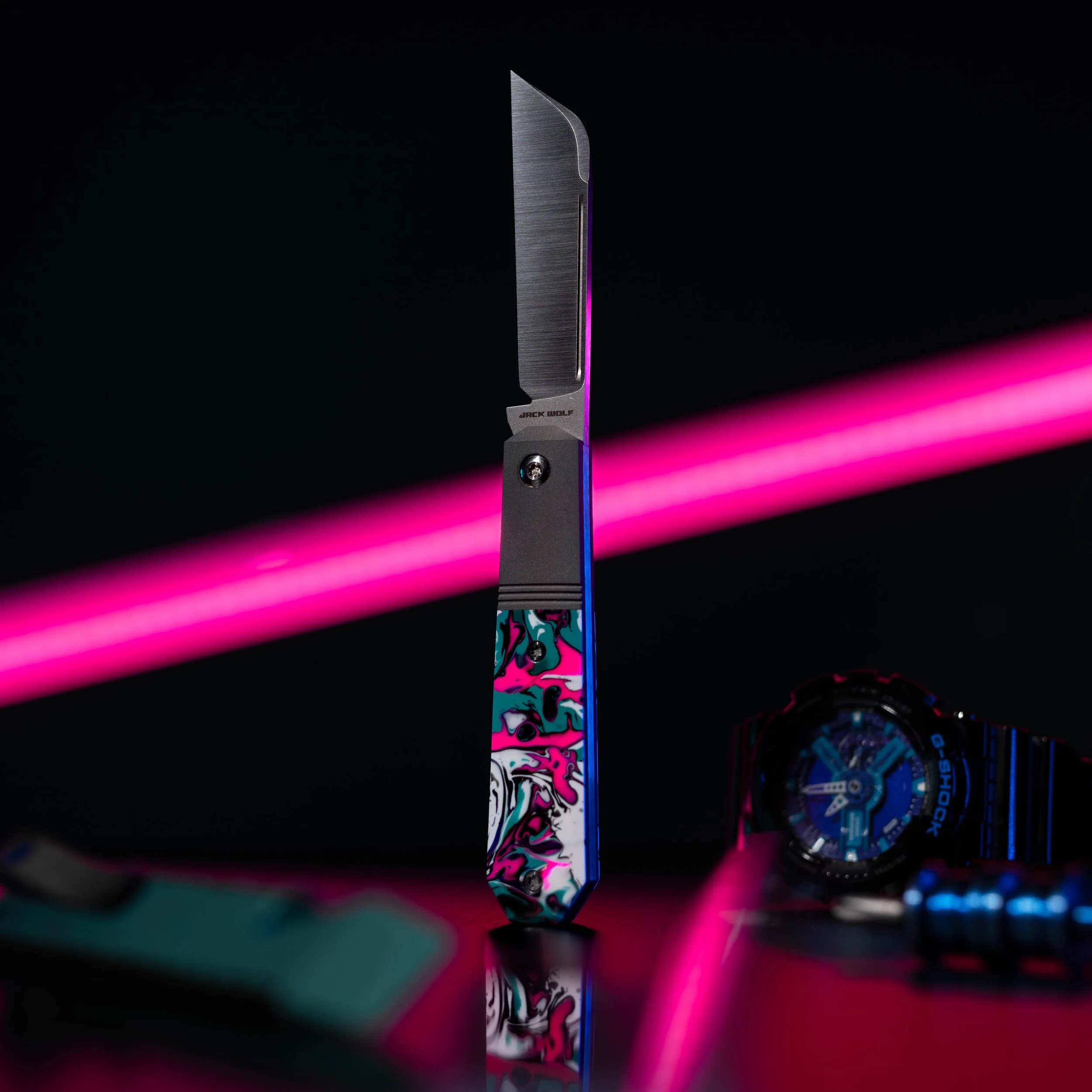 A folding knife with a colorful marbled handle and a partially open brushed metal blade, positioned upright with pink neon lights in the background, a watch and other objects partially visible on a reflective surface.