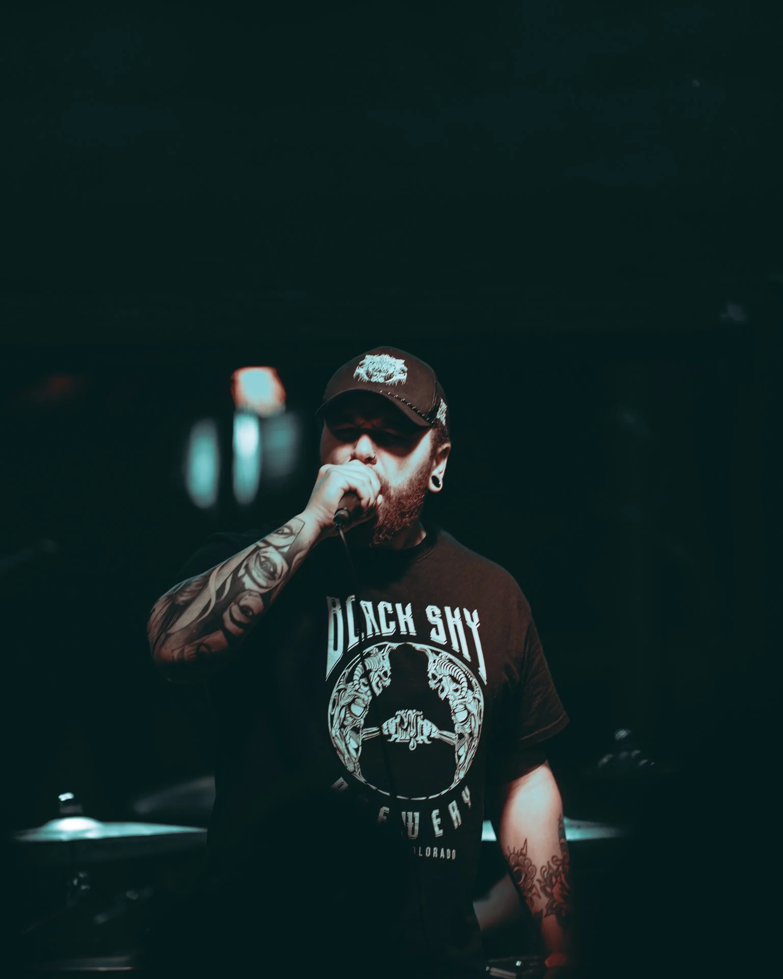 A man singing into a microphone, wearing a black cap and a black t-shirt with a graphic design and text, tattoos visible on his arms, in a dark setting with stage lights.
