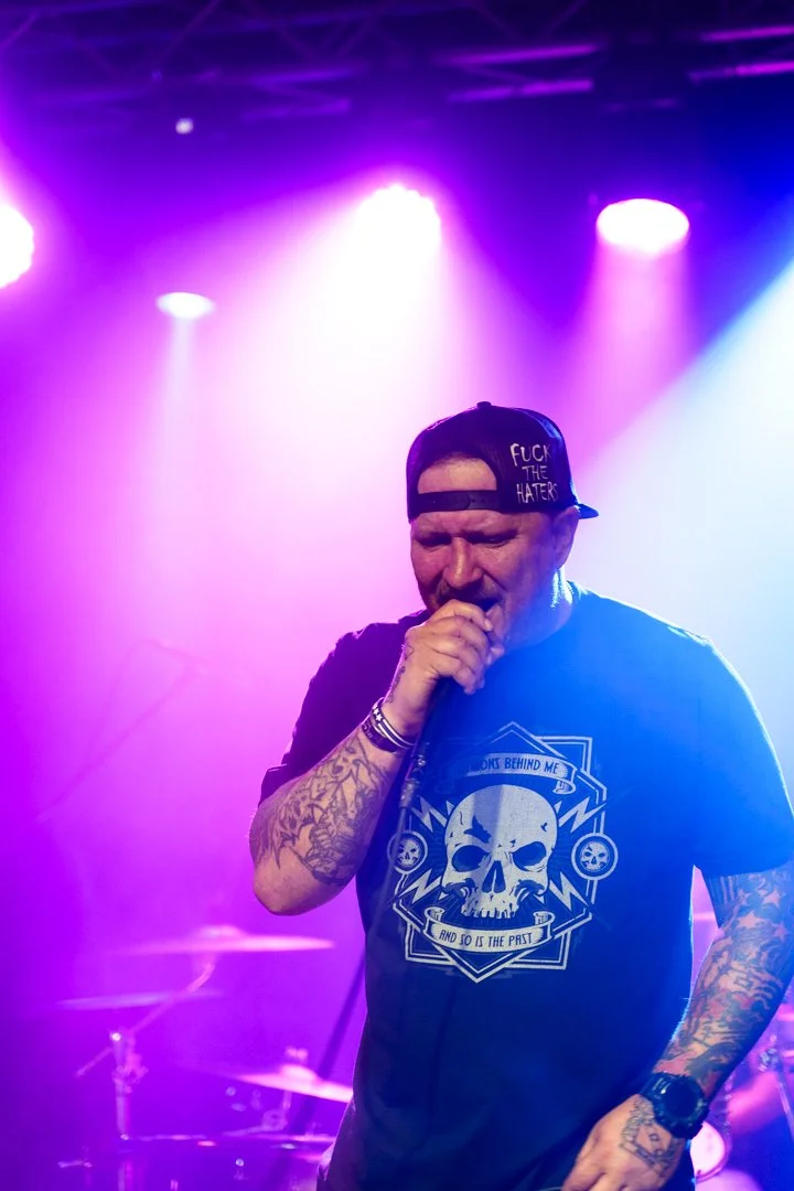 Male vocalist singing into microphone on stage with purple and blue lighting, wearing a black cap with the text "FUCK THE HATERS," a black t-shirt with a skull graphic, tattoos on arms, and a wristwatch.