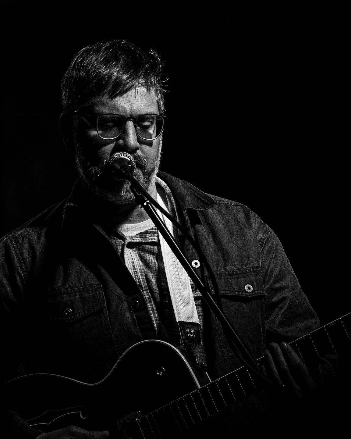 A man with glasses, a beard, and mustache playing an electric guitar and singing into a microphone in a dark setting.