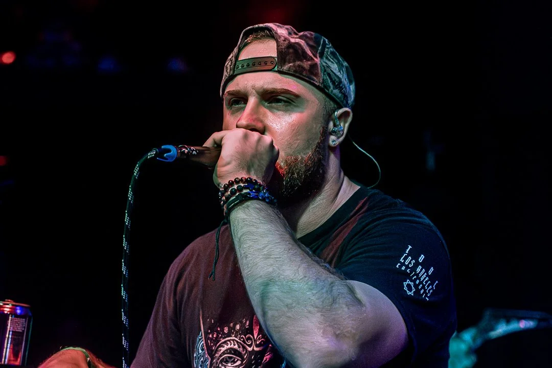 Man with a beard, wearing a cap backwards, singing into a microphone on stage, with colorful lighting.