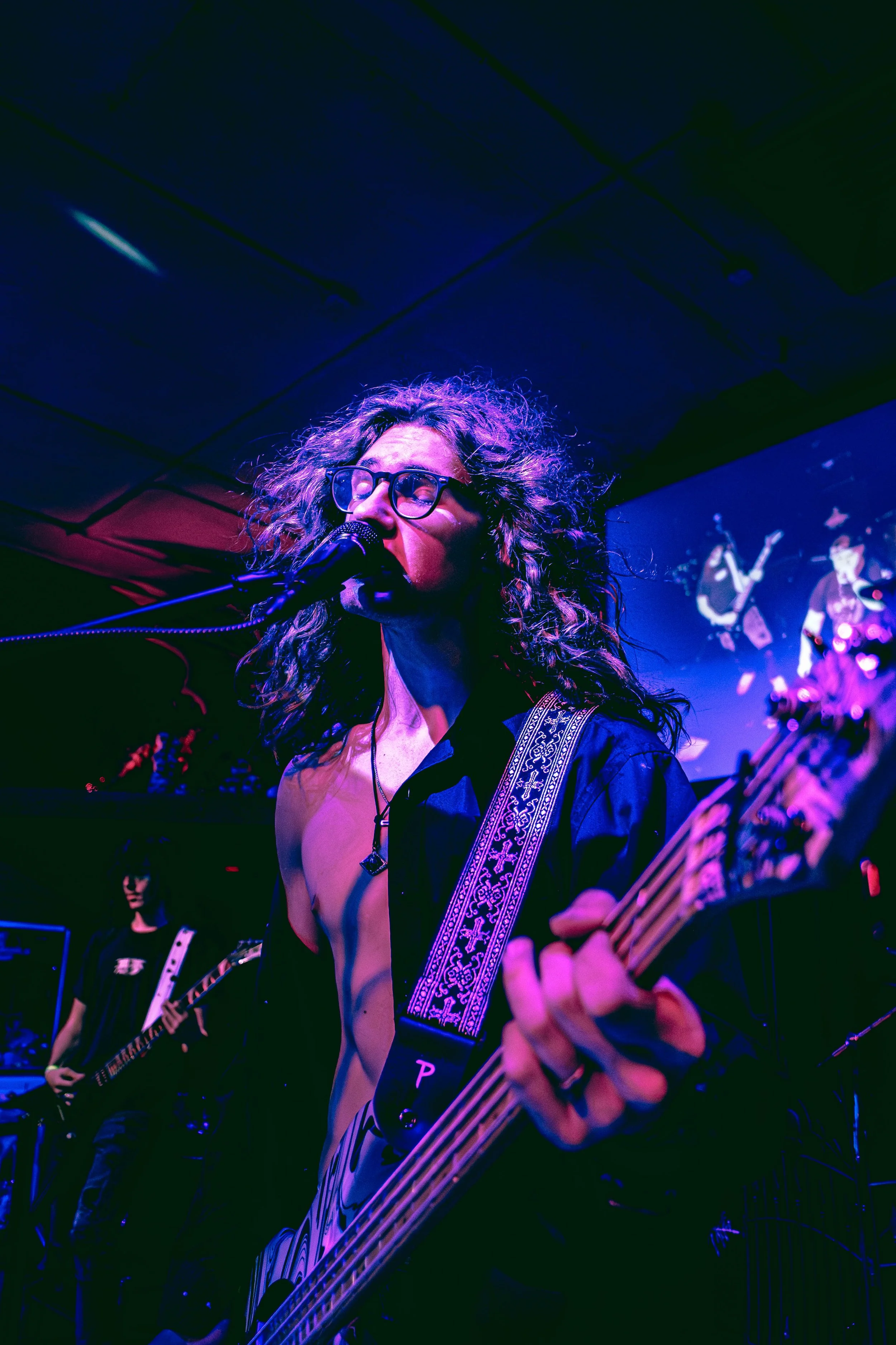 A male musician with curly hair blowing in the wind and glasses playing an electric bass guitar during a performance, with a microphone in front of him and a band in the background, under colorful stage lighting.