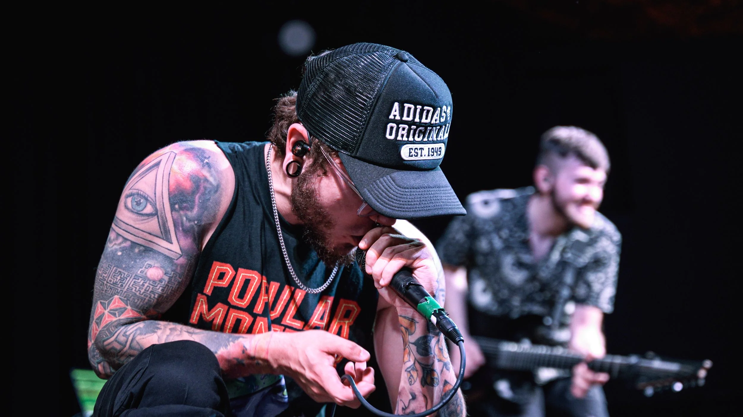 A man with tattoos and a baseball cap singing into a microphone, with a smiling man playing a guitar in the background.