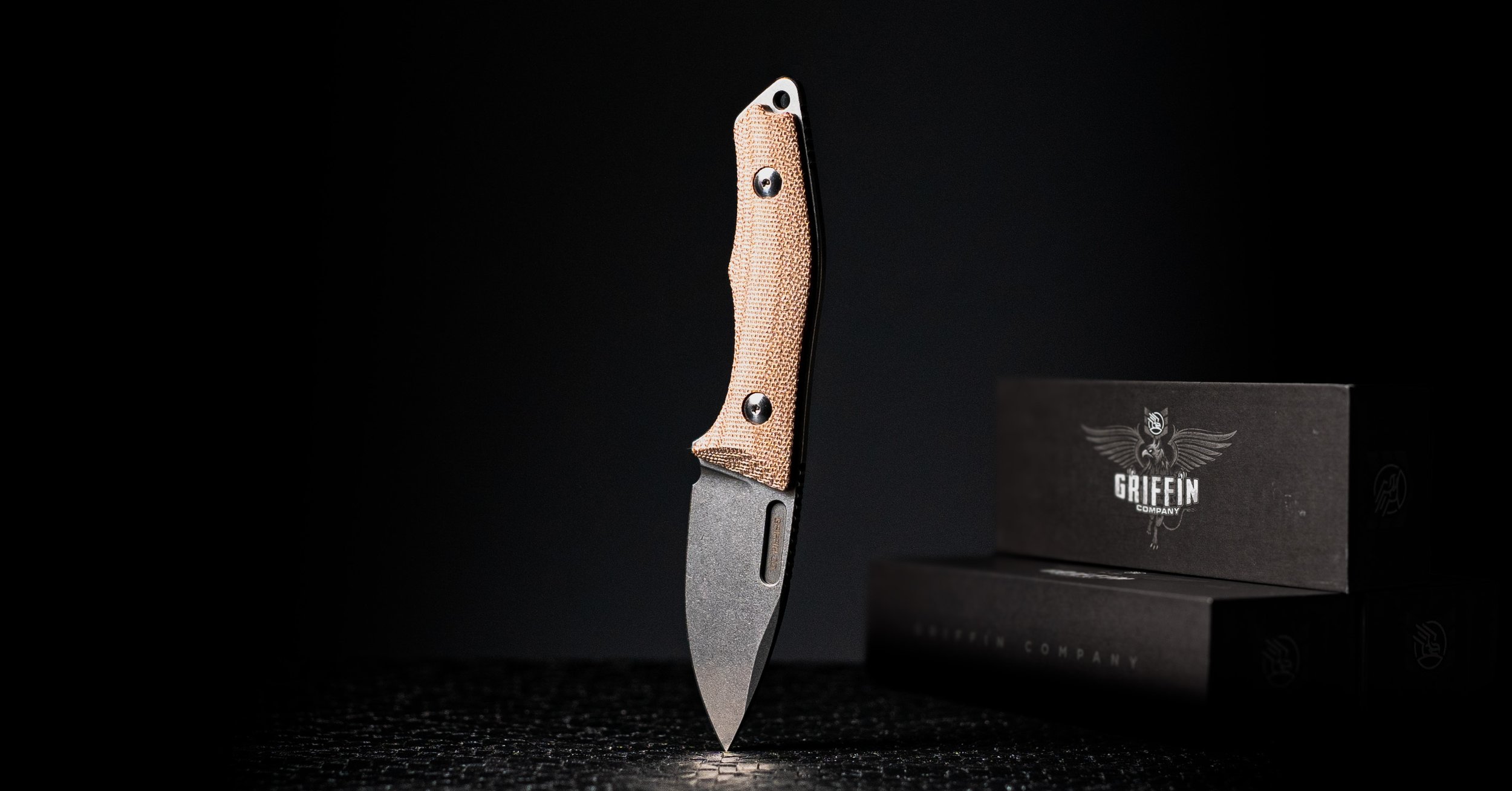 A tactical fixed blade knife with a natural micarta handle, standing upright on a dark surface, with a black box with a griffin logo and the words "Griffin Company" beside it.