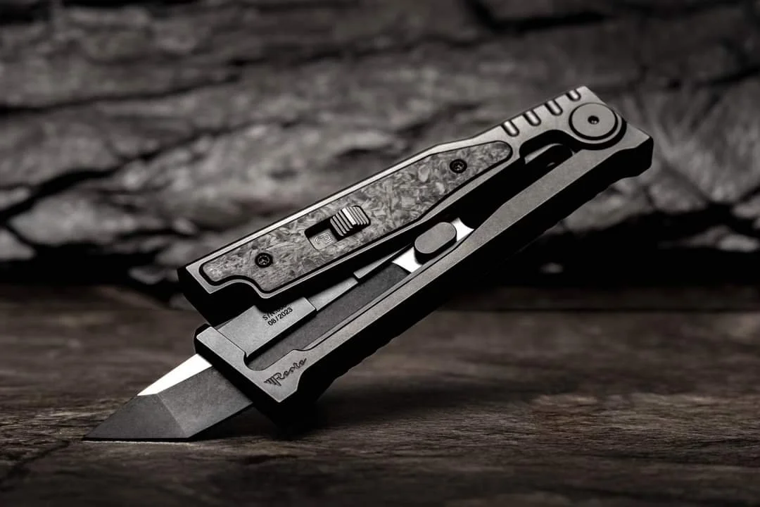 A gravity knife with a partially open blade, featuring a sleek black handle with textured grip and metallic accents, resting on a wooden surface with a rustic background.