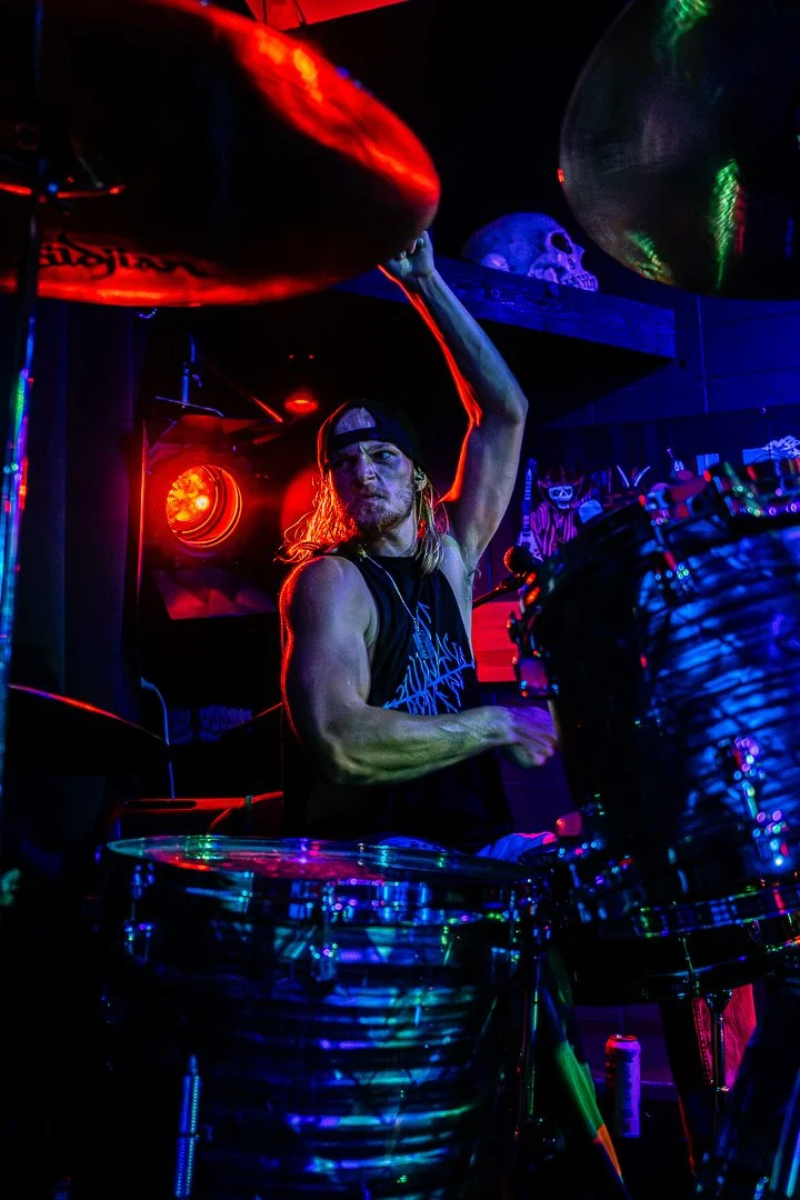 A drummer with long hair wearing a backwards baseball cap and a T-shirt, performing on stage under colorful neon lights, surrounded by drums and musical equipment.