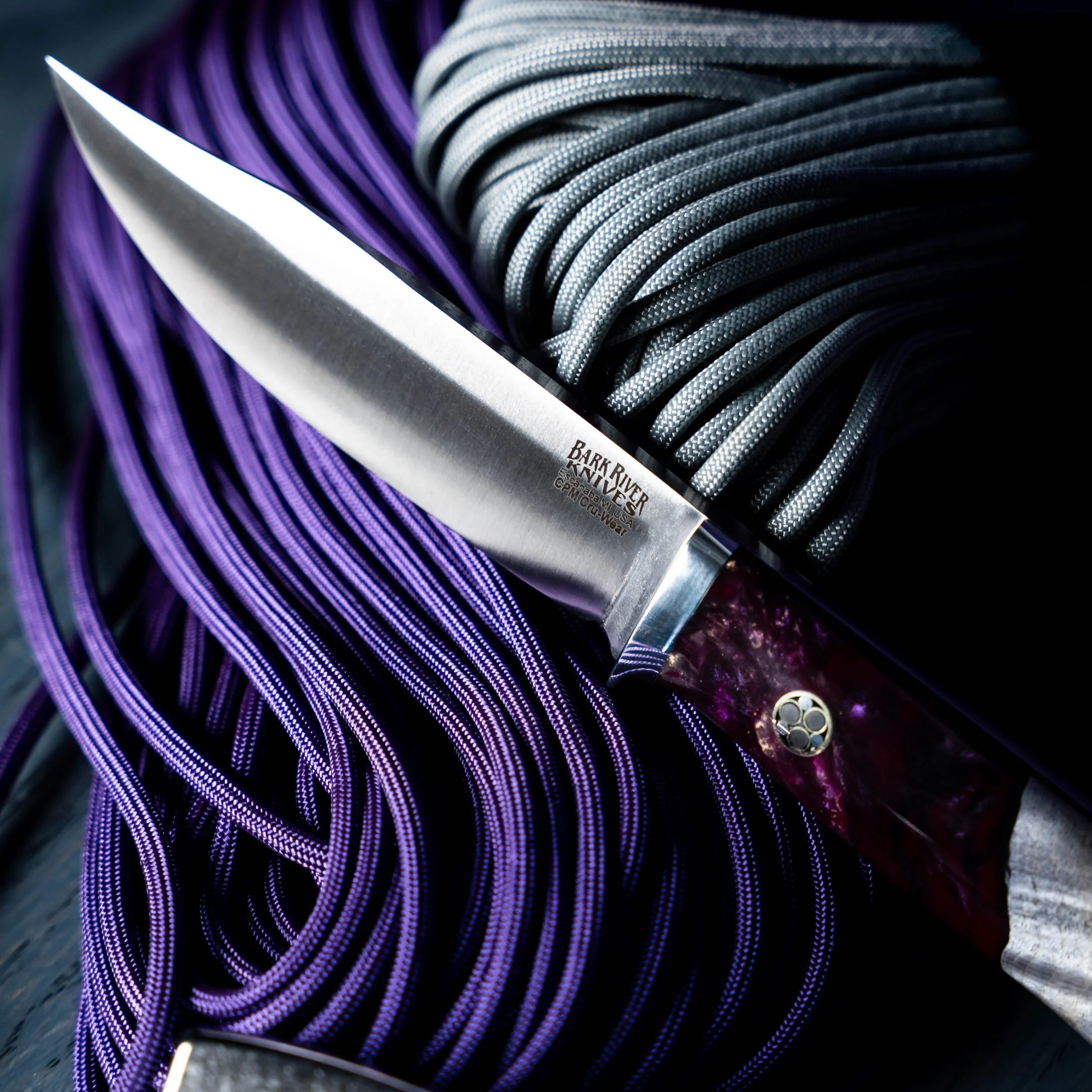 A kitchen knife with a purple resin and grey maple burl handle resting on bundles of purple and gray paracord.