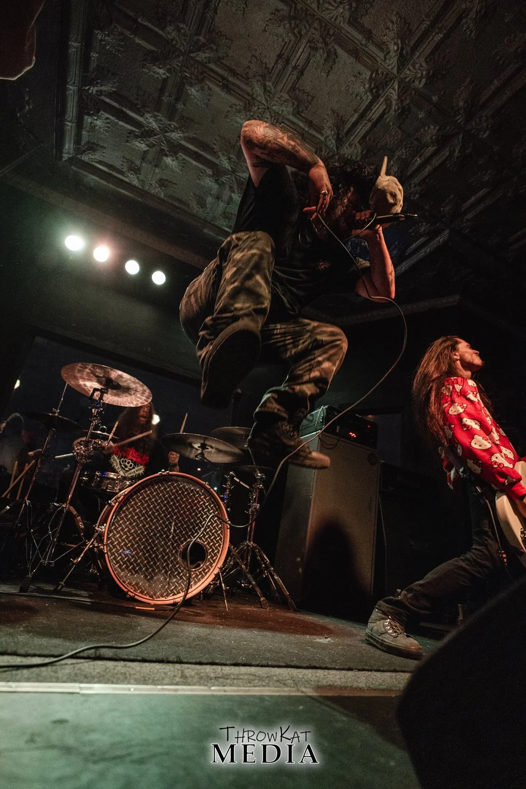 A band performs live on stage with a lead singer jumping in the air, holding a microphone. The stage includes a drummer in the background and a female guitarist to the right. The setting appears to be a dark, intimate music venue with a decorative ce