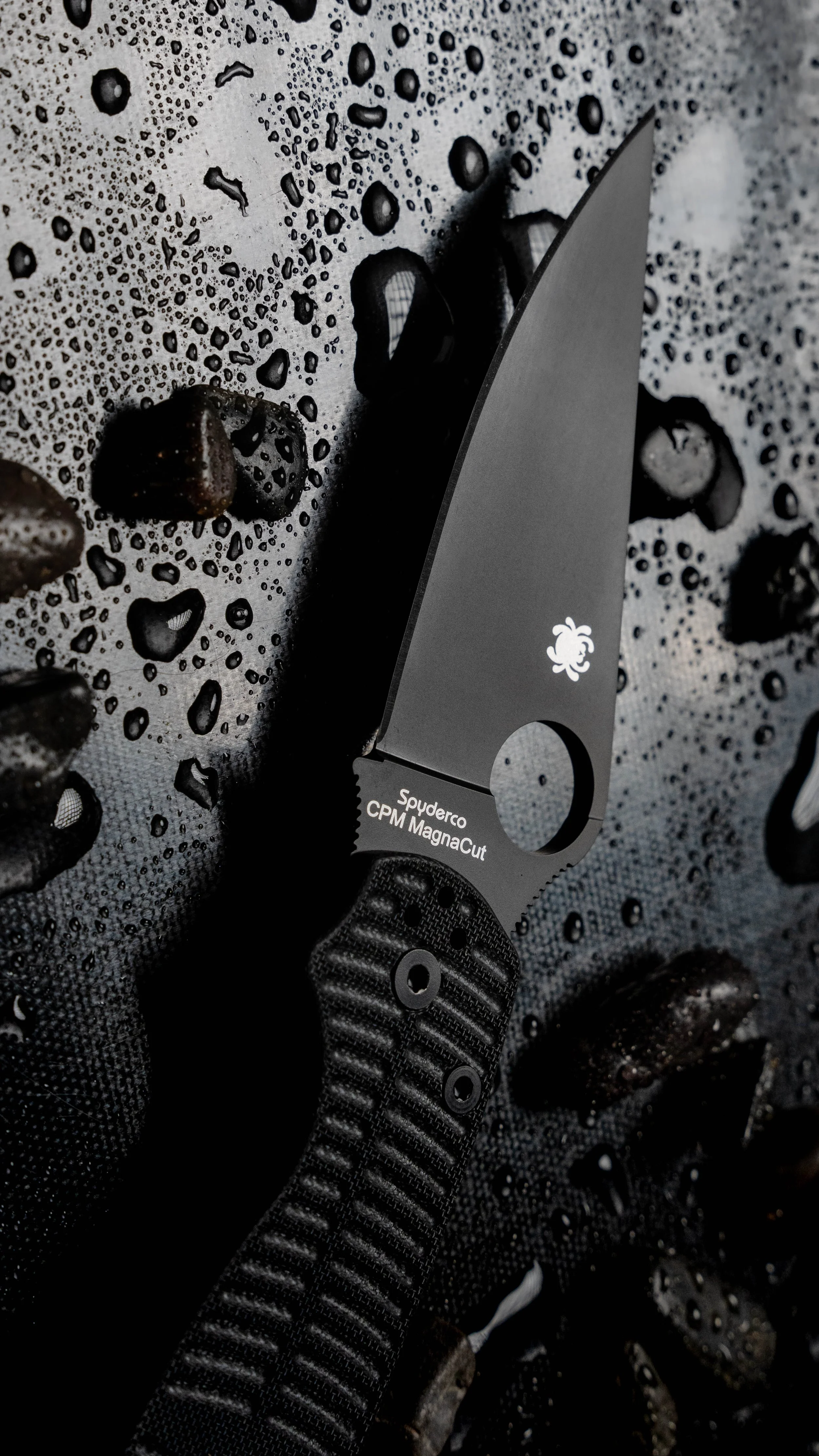 Close-up of a black Spyderco CPM Magnacut folding knife with a textured handle, placed on a wet, pebbled surface with water droplets and dark rocks.